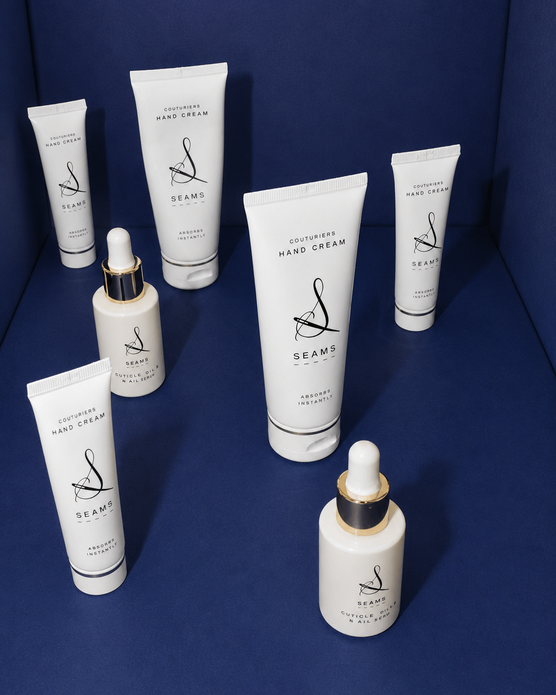 SEAMS hand cream and cuticle oil products displayed on a rich blue background in a luxury beauty flat lay. White packaging with black typography and gold accents creates a sophisticated, premium skincare presentation.