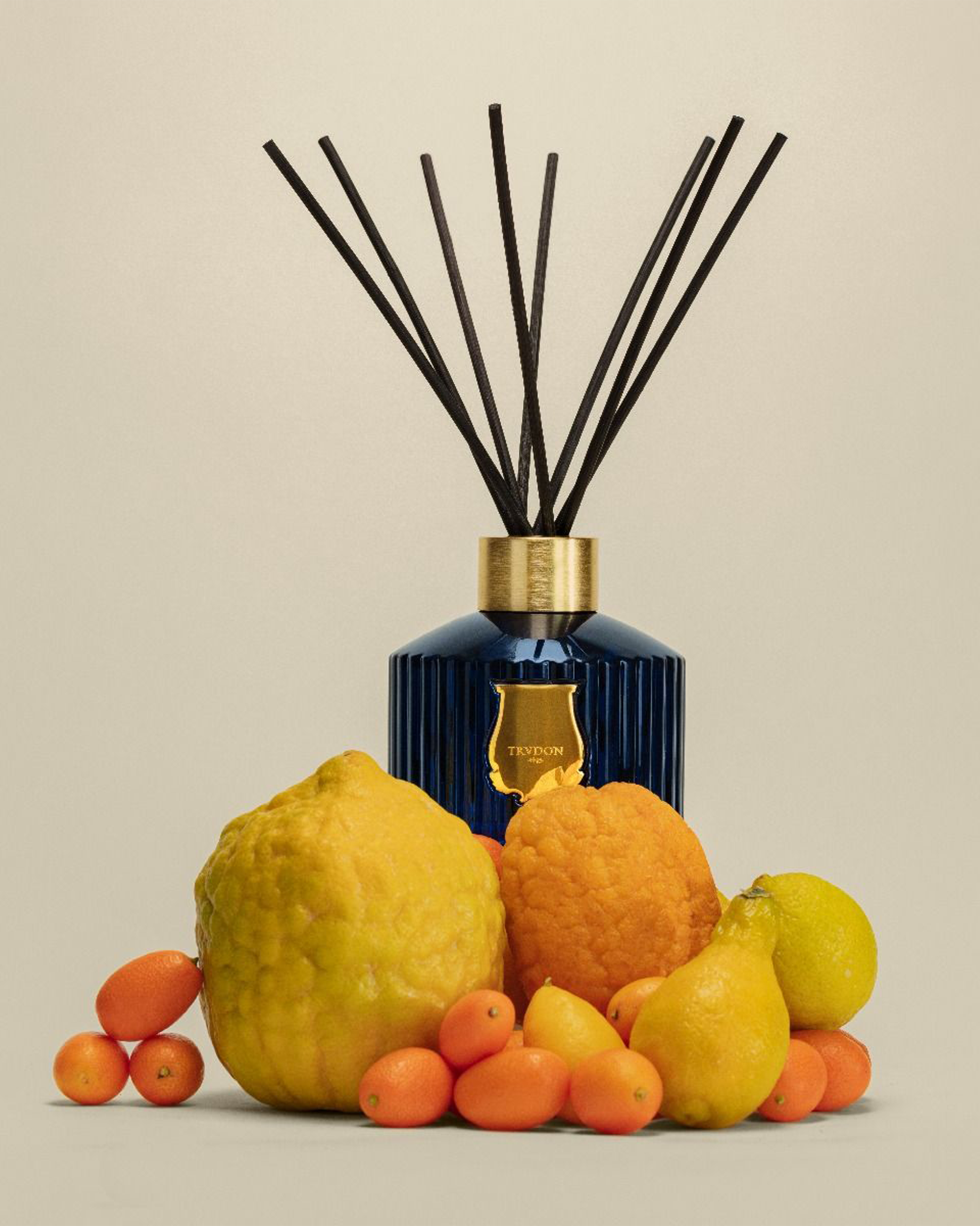 A luxury reed diffuser with a ribbed cobalt blue glass bottle, gold collar, and gold crest emblem, surrounded by fresh citrus fruits including a large lemon, oranges, and pears, with black reeds fanned out against a warm beige background.