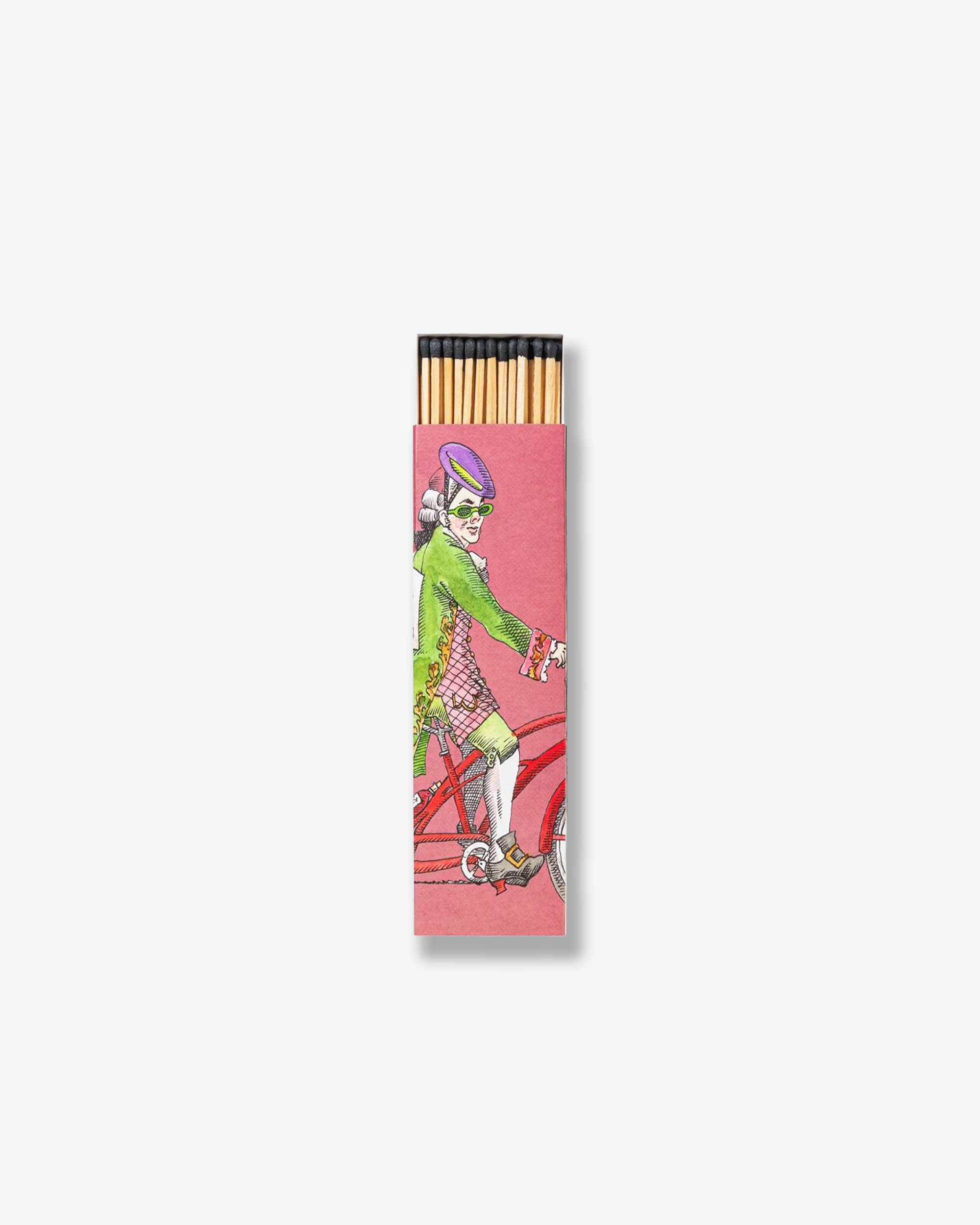 A pink Trudon matchbox decorated with a vintage-style illustration of an 18th-century man wearing green glasses, a purple cap, and a green coat, riding a red bicycle. Black-tipped match heads emerge from the top opening of the box.
