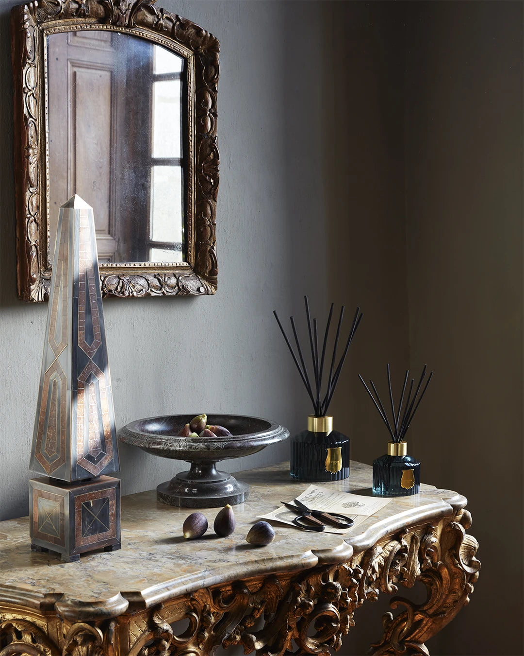 Two Trudon reed diffusers of different sizes displayed on an ornate marble console table alongside fresh figs, an obelisk, a stone bowl, and a vintage mirror.