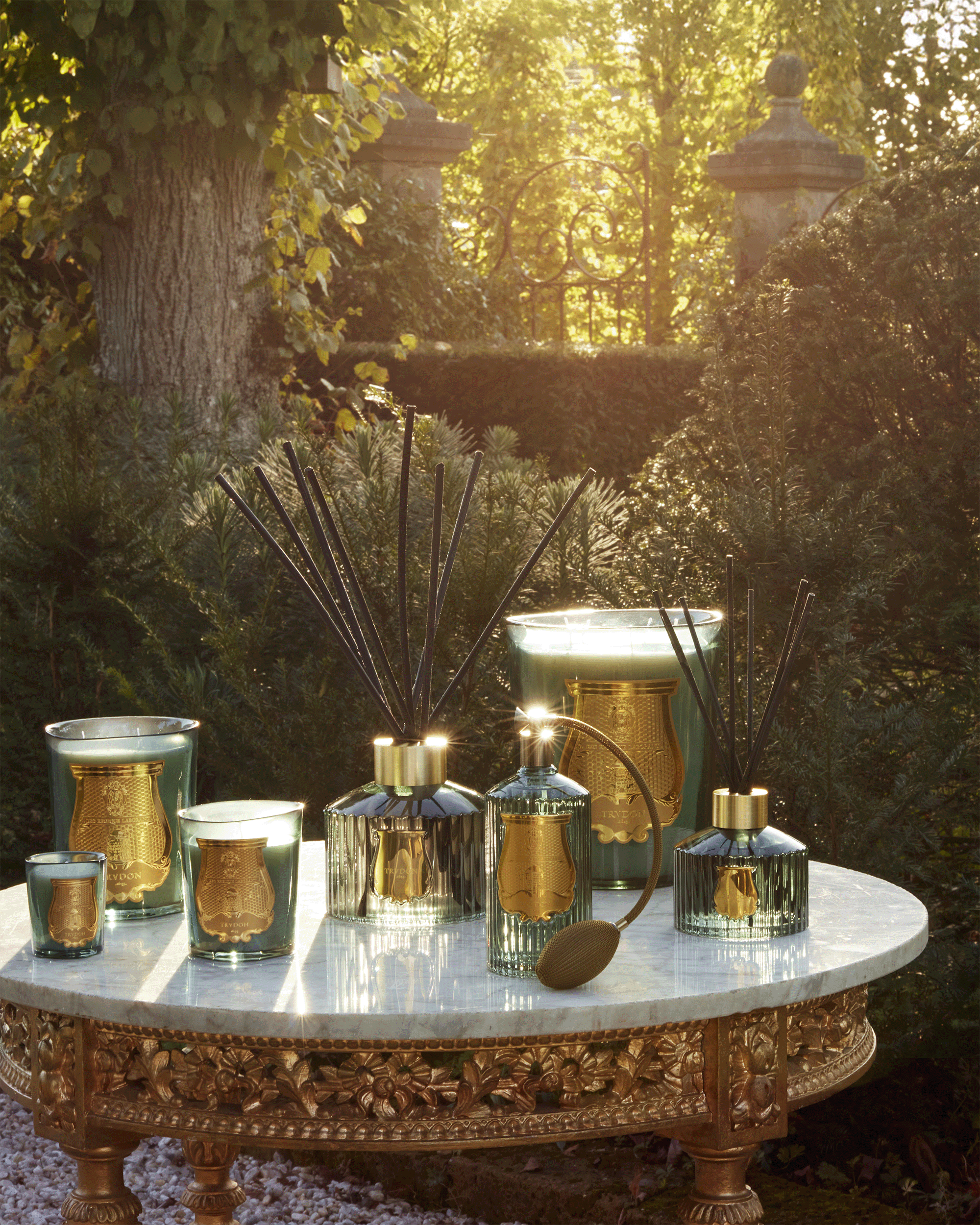 A collection of decorative home fragrance items, including reed diffusers and candles in gold and smoky glass, arranged on an ornate round table outdoors in a garden setting with warm sunlight filtering through trees.