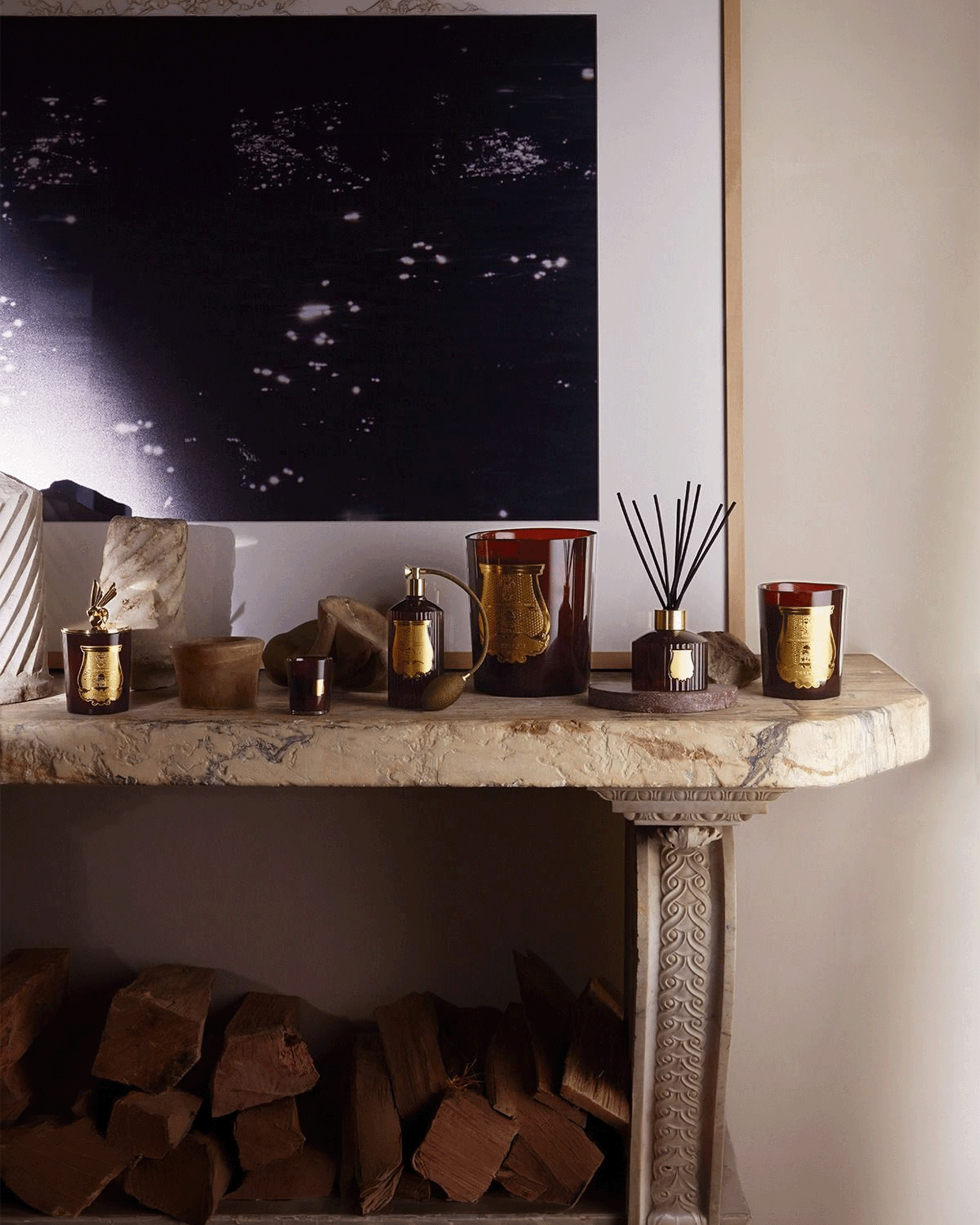 A styled interior scene showing a marble console table with home decor items, including amber glass candle jars, a reed diffuser, and a metallic vessel, set against a dark abstract wall art and wood logs below.