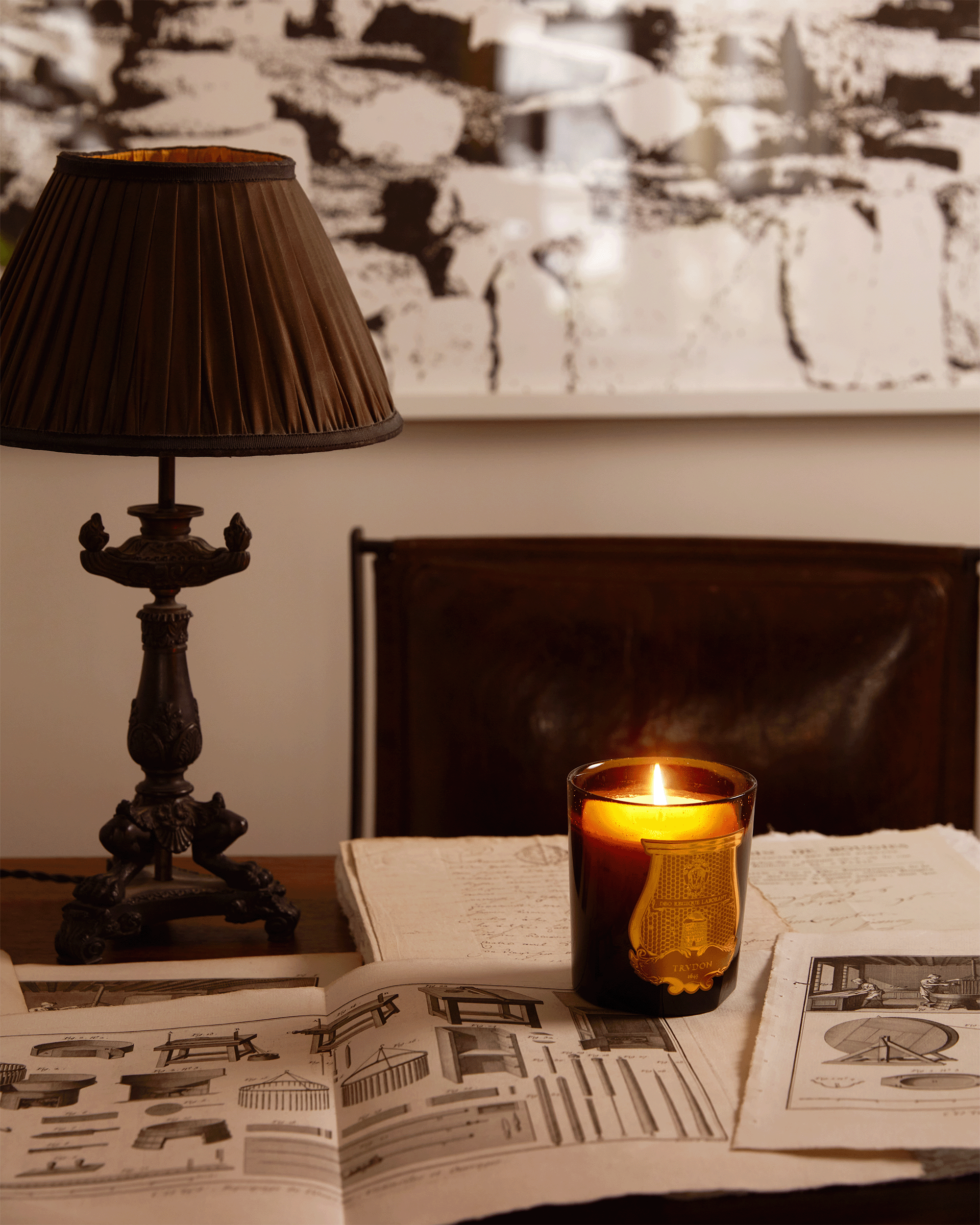 A lit amber glass candle with a gold crest sits on an open book on a table, alongside a dark wood table lamp and architectural print pages, with a dark leather chair and abstract wall art in the background.