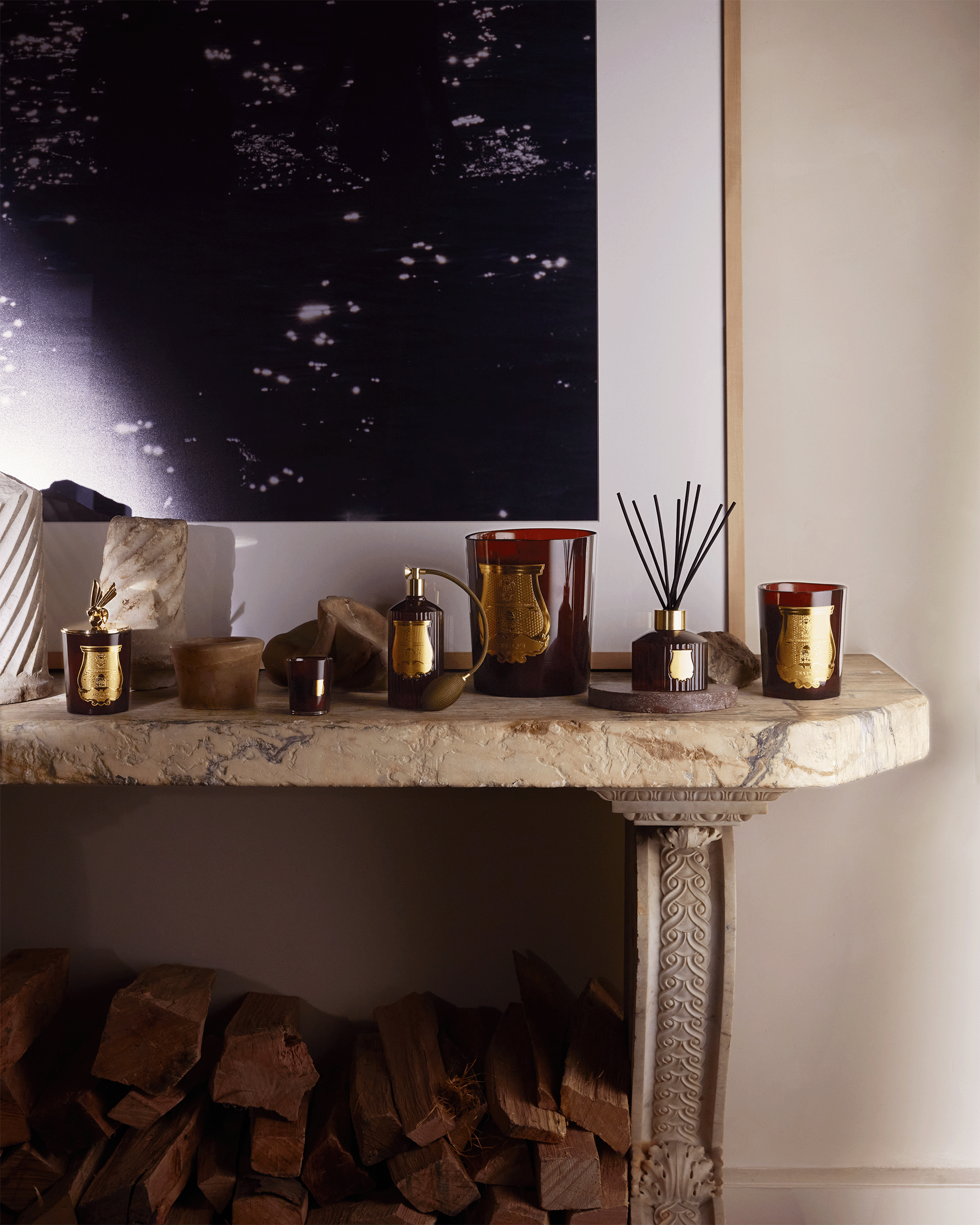 A styled interior scene showing a marble console table holding several amber glass candle jars, a reed diffuser, and small decorative vessels, with stacked firewood beneath and a dark abstract wall art piece above.