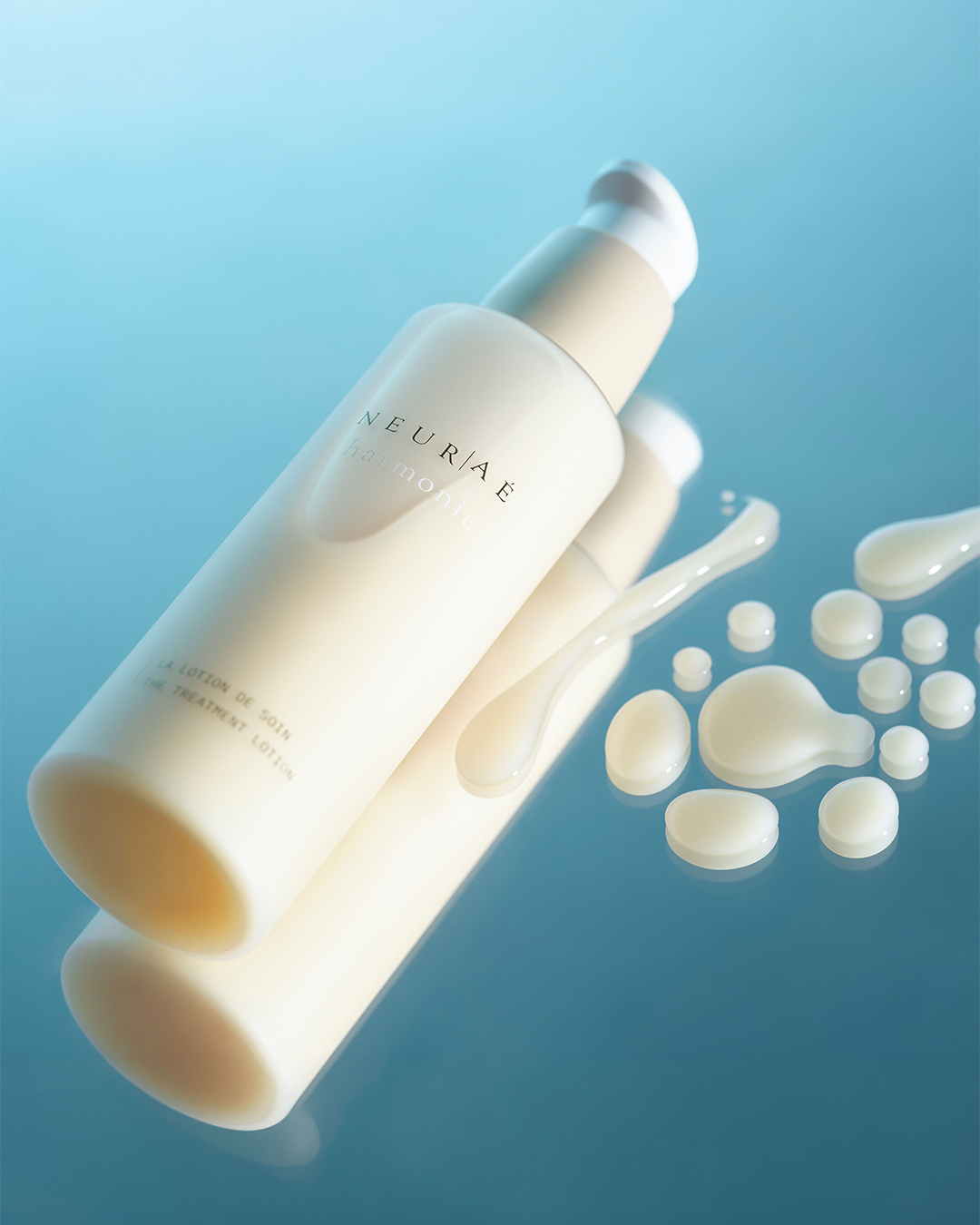 The Neuraé Harmonie Treatment Lotion bottle lying on a reflective blue surface next to scattered droplets of its liquid texture.