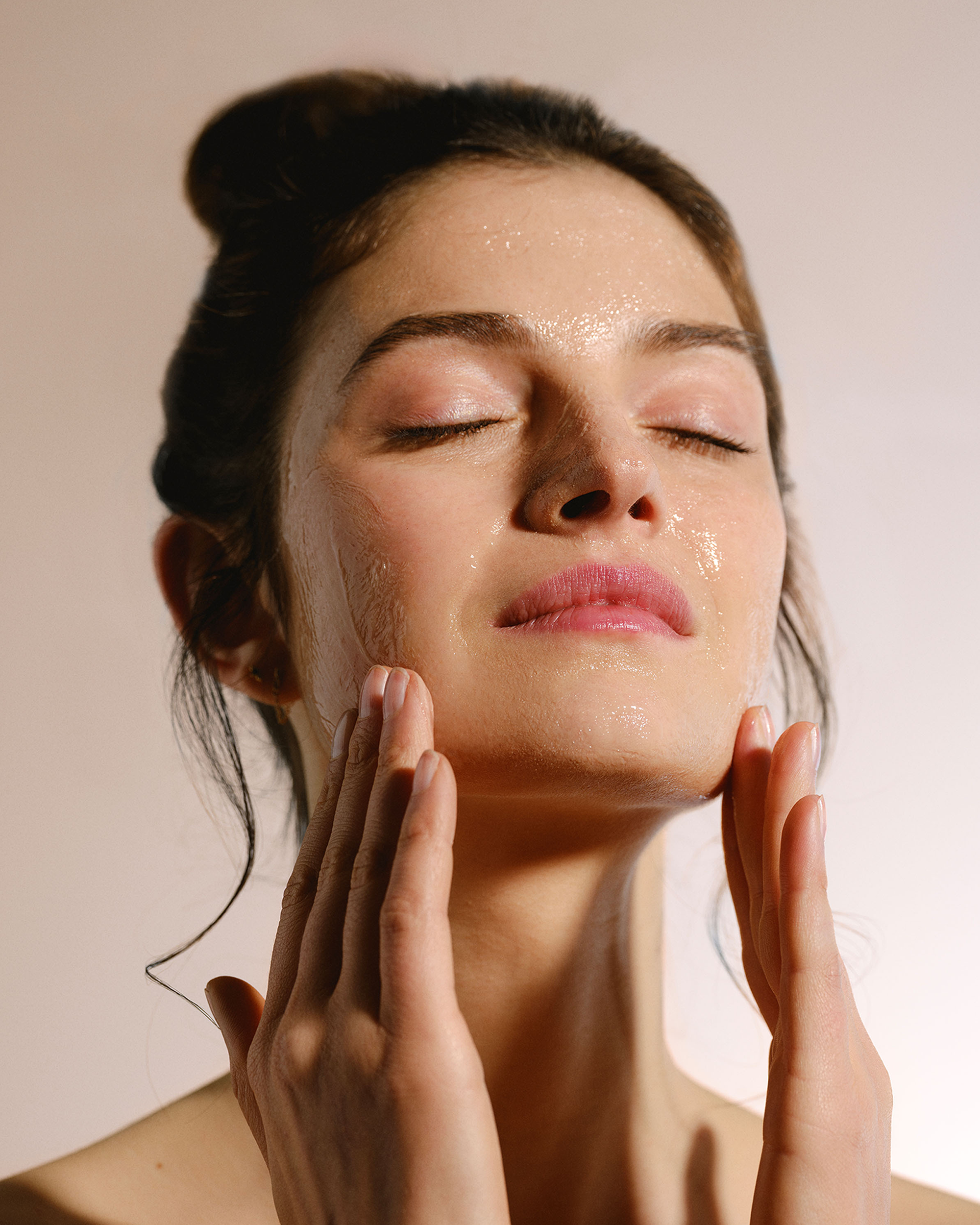 A beauty lifestyle portrait of a woman with her eyes closed, gently massaging a clear gel cleanser onto her glowing face.