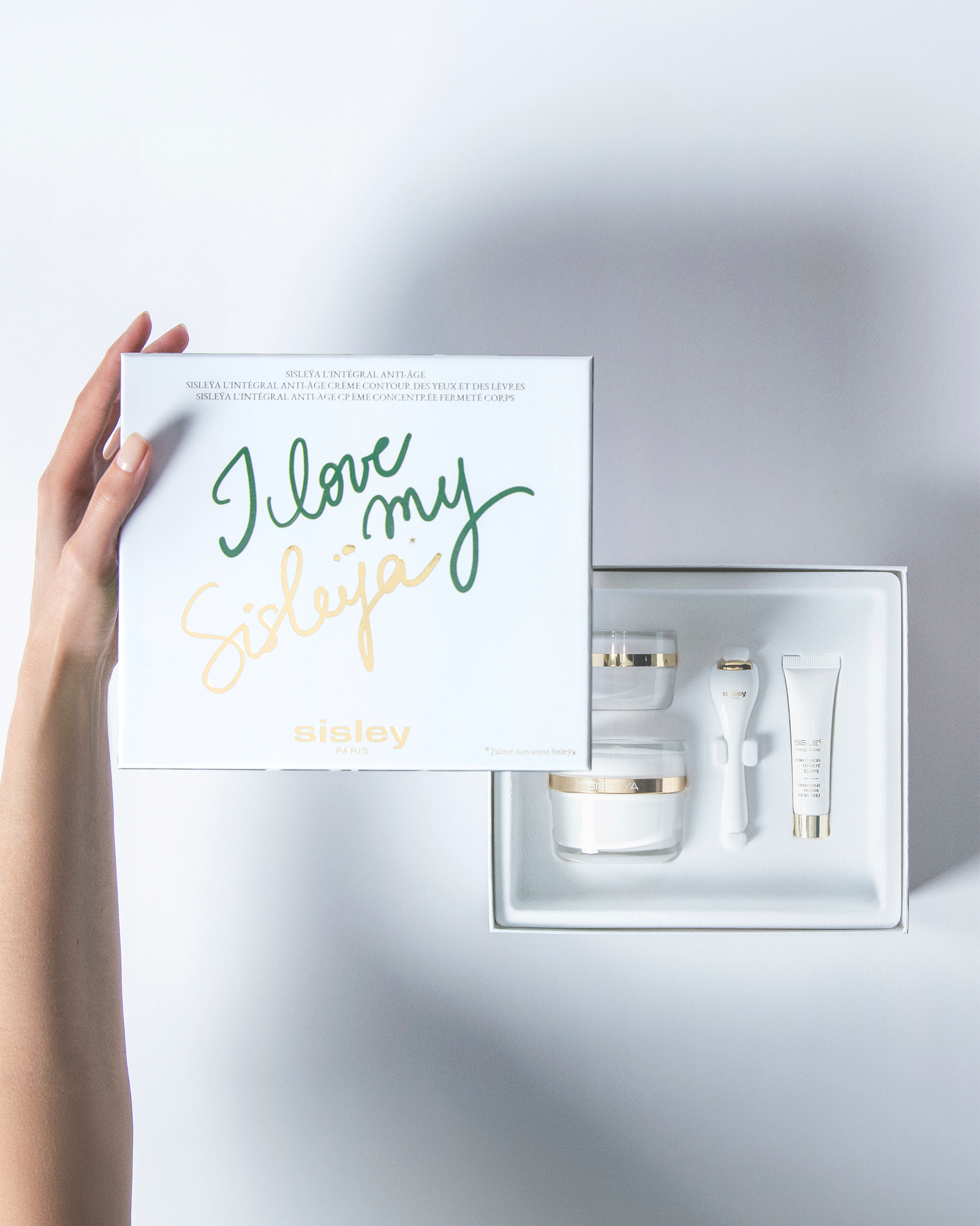 A hand holds a white Sisley gift box with the text 'I love my Sisley' in green and gold script, next to an open tray containing a skincare set with white and gold containers and tools, on a light gray background.