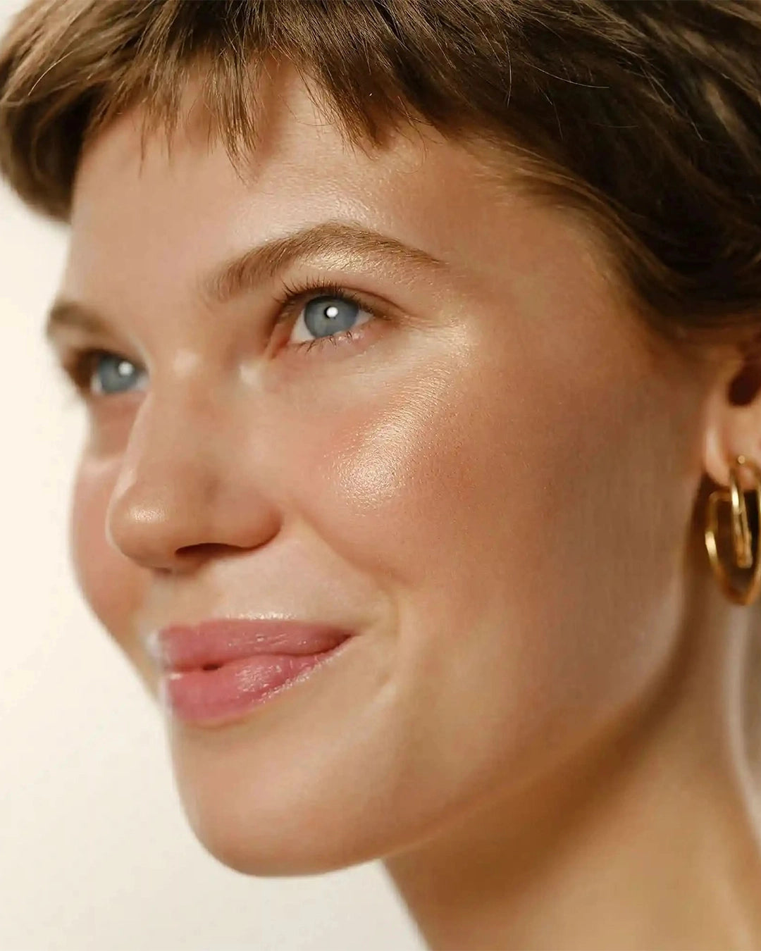 Close-up portrait of a woman with short brown hair, clear blue eyes, and radiant, sun-kissed dewy skin looking off-camera.