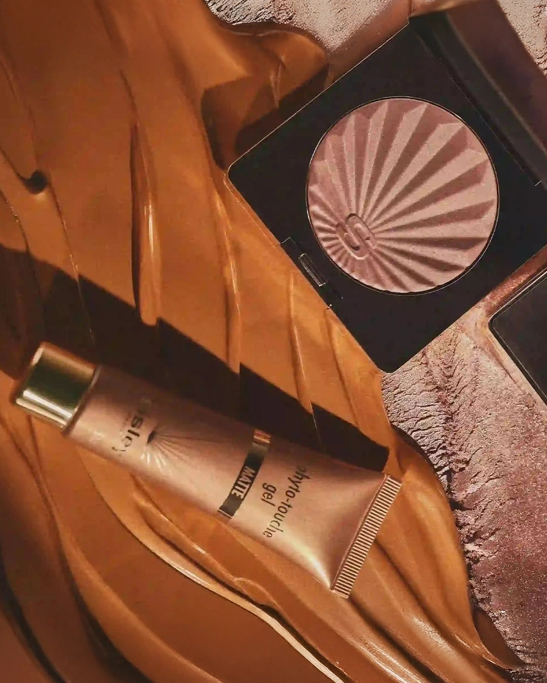 An aesthetic, stylized flat lay featuring the Sisley Phyto-Touche Gel Matte tube and an open compact powder resting on a glossy backdrop of swirled bronze liquid gel.