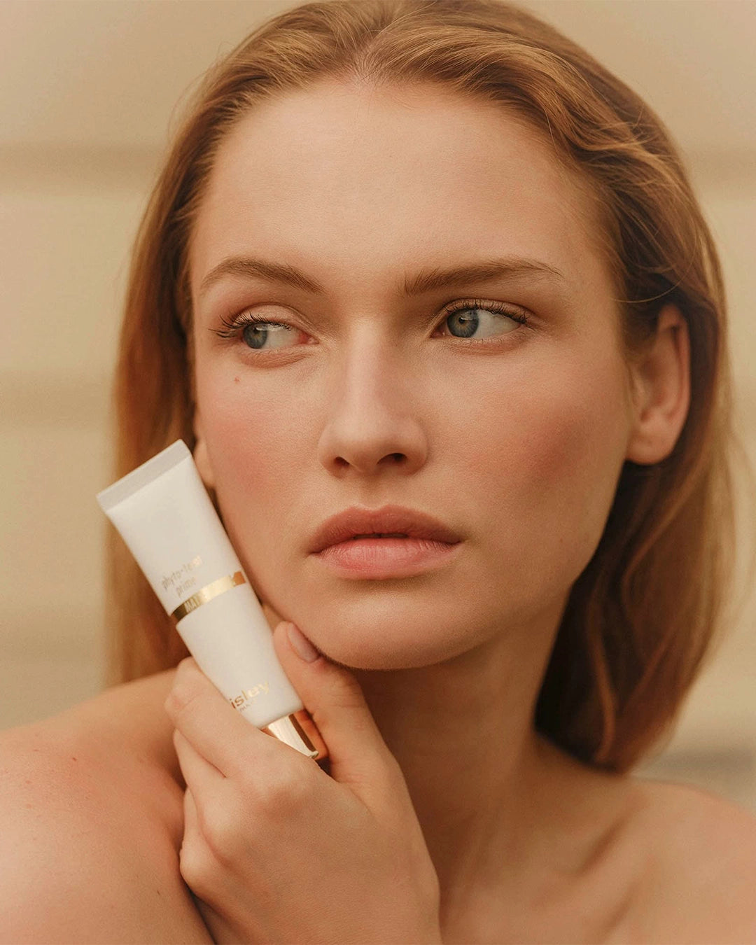 Close-up portrait of a woman with auburn hair resting her chin against a white tube of Sisley Phyto-Teint Primer.