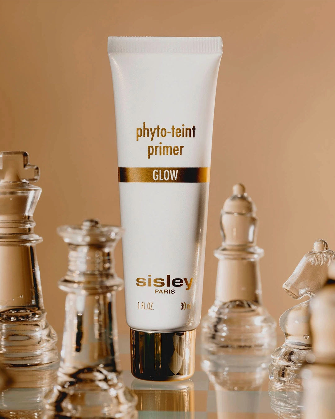A white squeeze tube of Sisley Phyto-Teint Primer Glow standing vertically amidst frosted glass chess pieces on a reflective board.