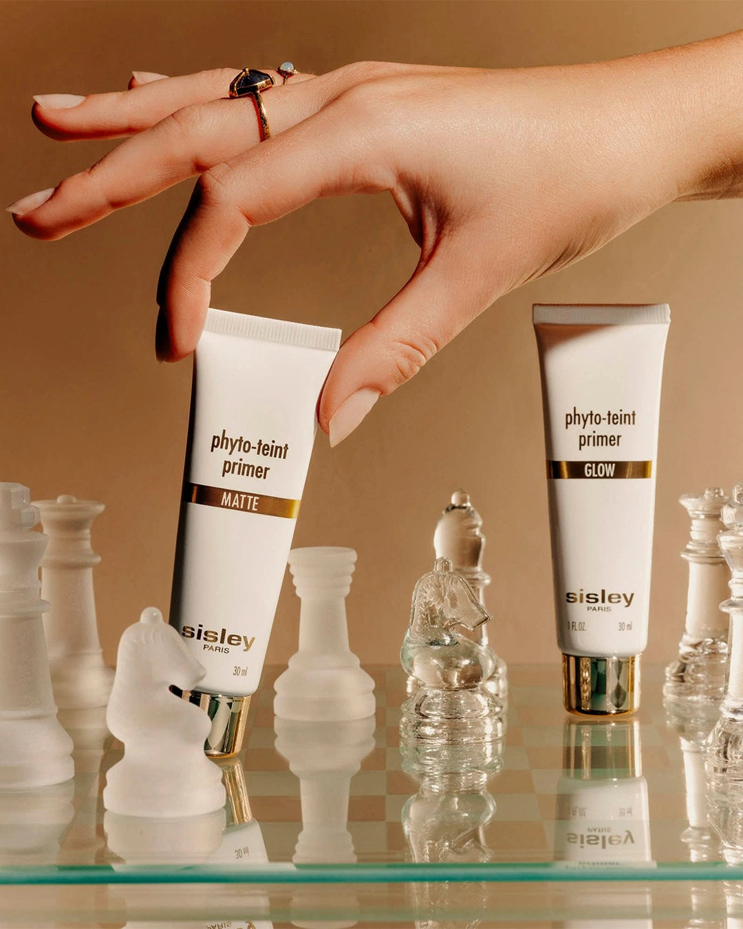 A hand wearing gold rings reaches down to pick up a tube of Sisley Phyto-Teint Primer Matte from a glass chessboard.