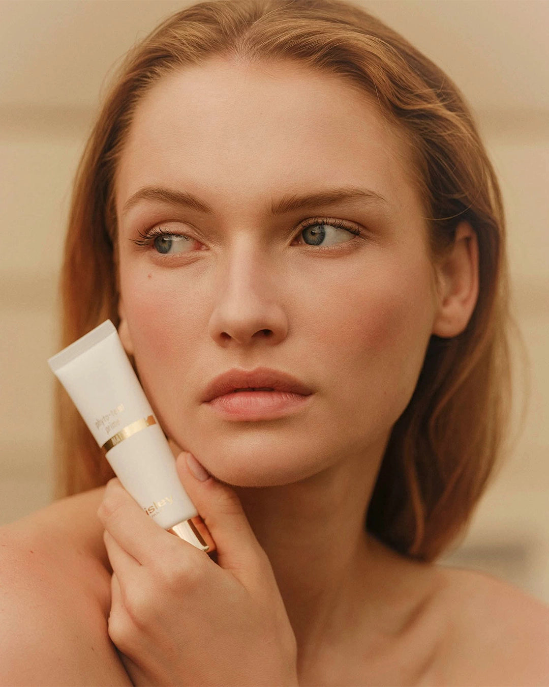 Close-up portrait of a woman with auburn hair resting her chin against a white tube of Sisley Phyto-Teint Primer.
