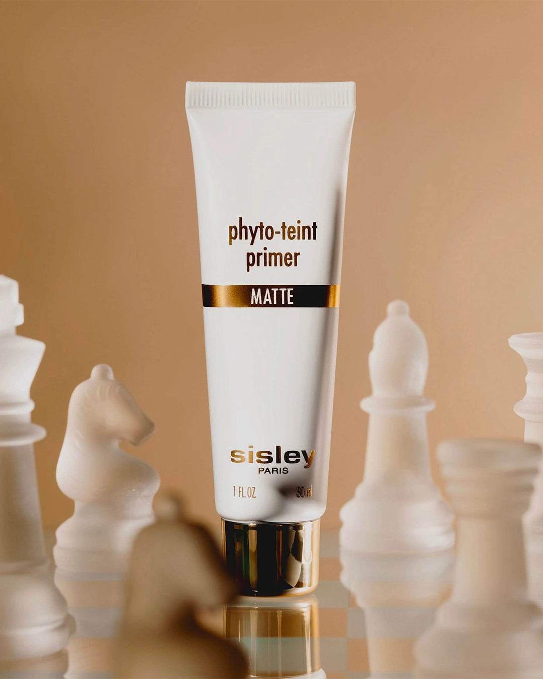 A white squeeze tube of Sisley Phyto-Teint Primer Matte standing vertically amidst frosted glass chess pieces on a reflective board.