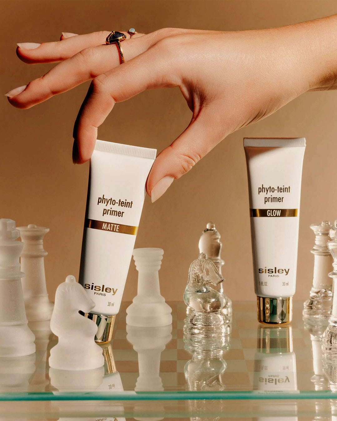 A hand wearing gold rings reaches down to pick up a tube of Sisley Phyto-Teint Primer Matte from a glass chessboard.