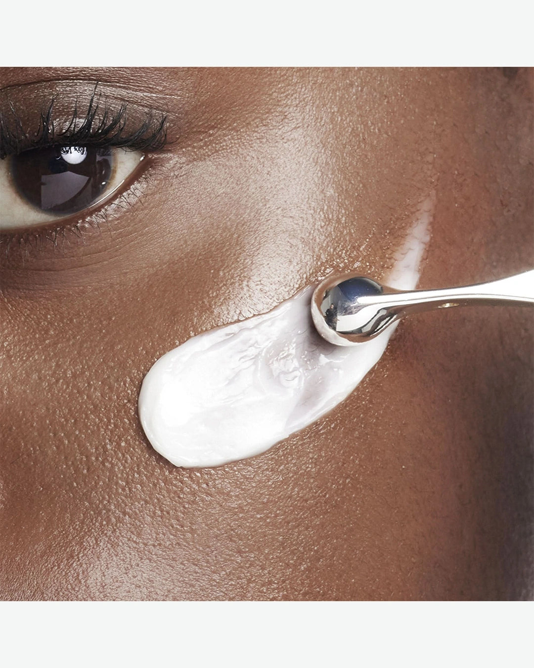 Close-up of a person applying white cream to their face with a silver spoon.