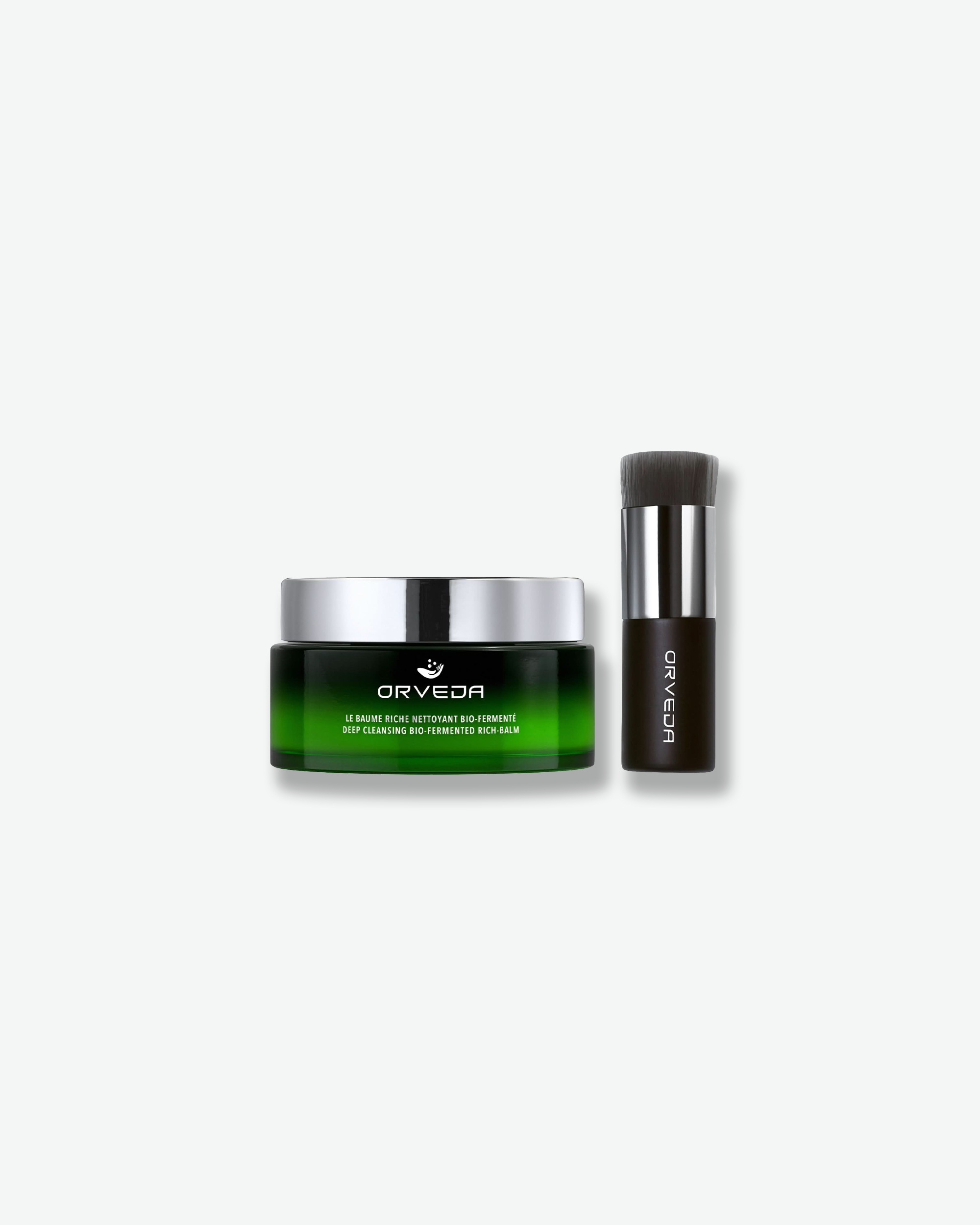Product shot featuring an Orveda "Deep Cleansing Bio-Fermented Rich-Balm" jar standing alongside a blending brush against a plain gray background. The short, wide green gradient jar with a silver cap sits next to a thick skincare brush featuring a dark handle, silver ferrule, and densely packed dark grey bristles.