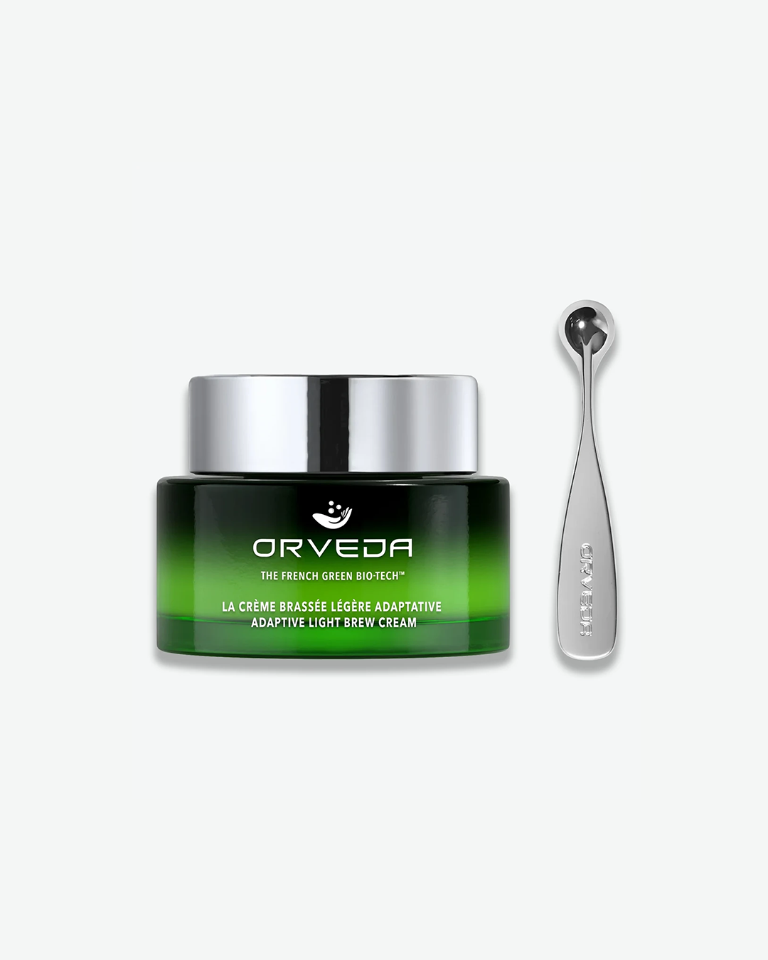 A product shot of an Orveda Adaptive Light Brew Cream jar next to its applicator tool, centered against a plain light gray background. The shallow glass jar features a dark green gradient that showcases a vibrant lime-green cream inside, sealed with a polished chrome silver cap. The front text reads "LA CRÈME BRASSÉE LÉGÈRE ADAPTATIVE ADAPTIVE LIGHT BREW CREAM". To its right stands a matching, vertical polished silver massage tool with a spherical tip.