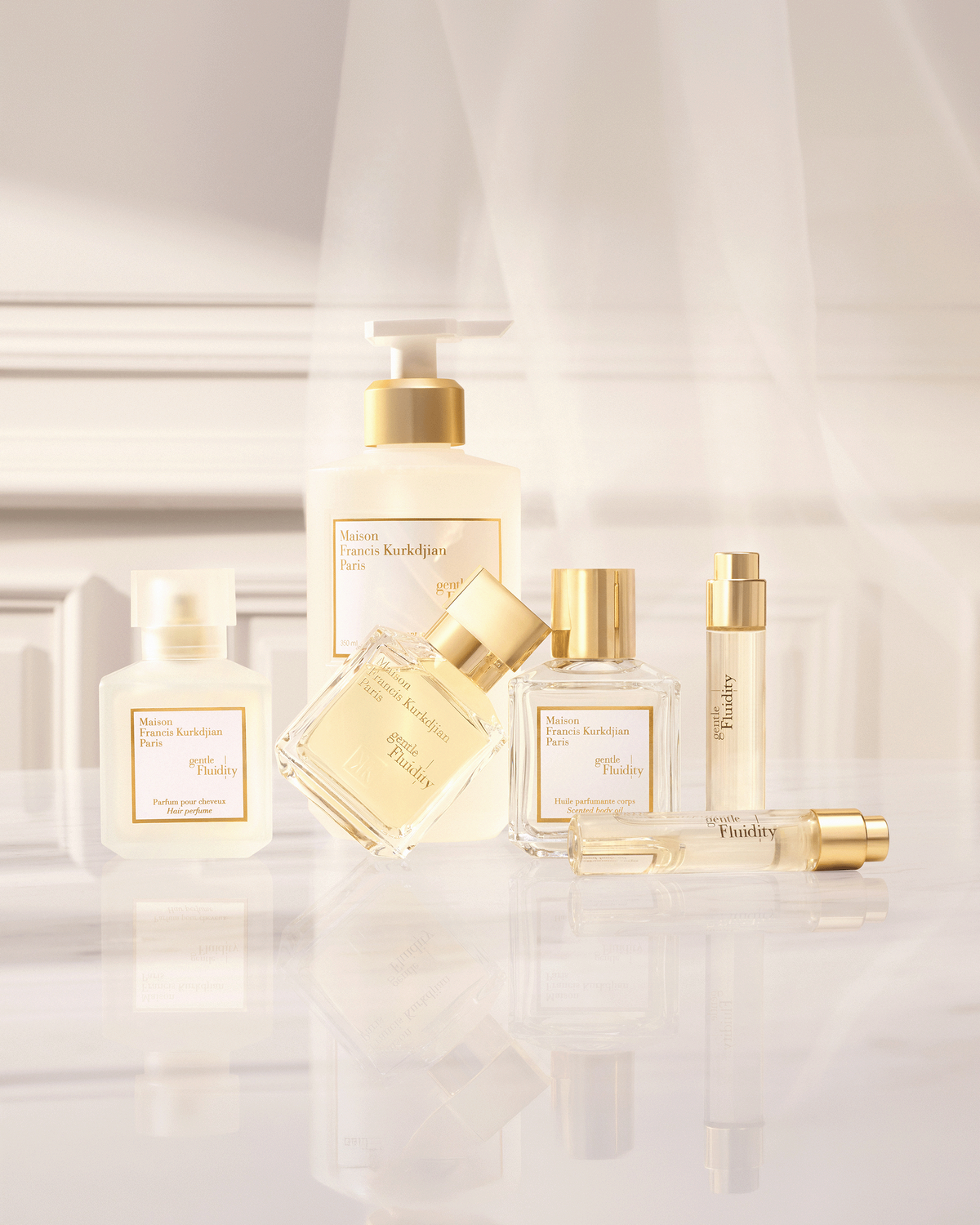 Maison Francis Kurkdjian Gentle Fluidity collection arranged on a reflective surface in a softly lit cream interior, featuring fragrance, body oil, hair perfume, cleansing gel, and travel formats.