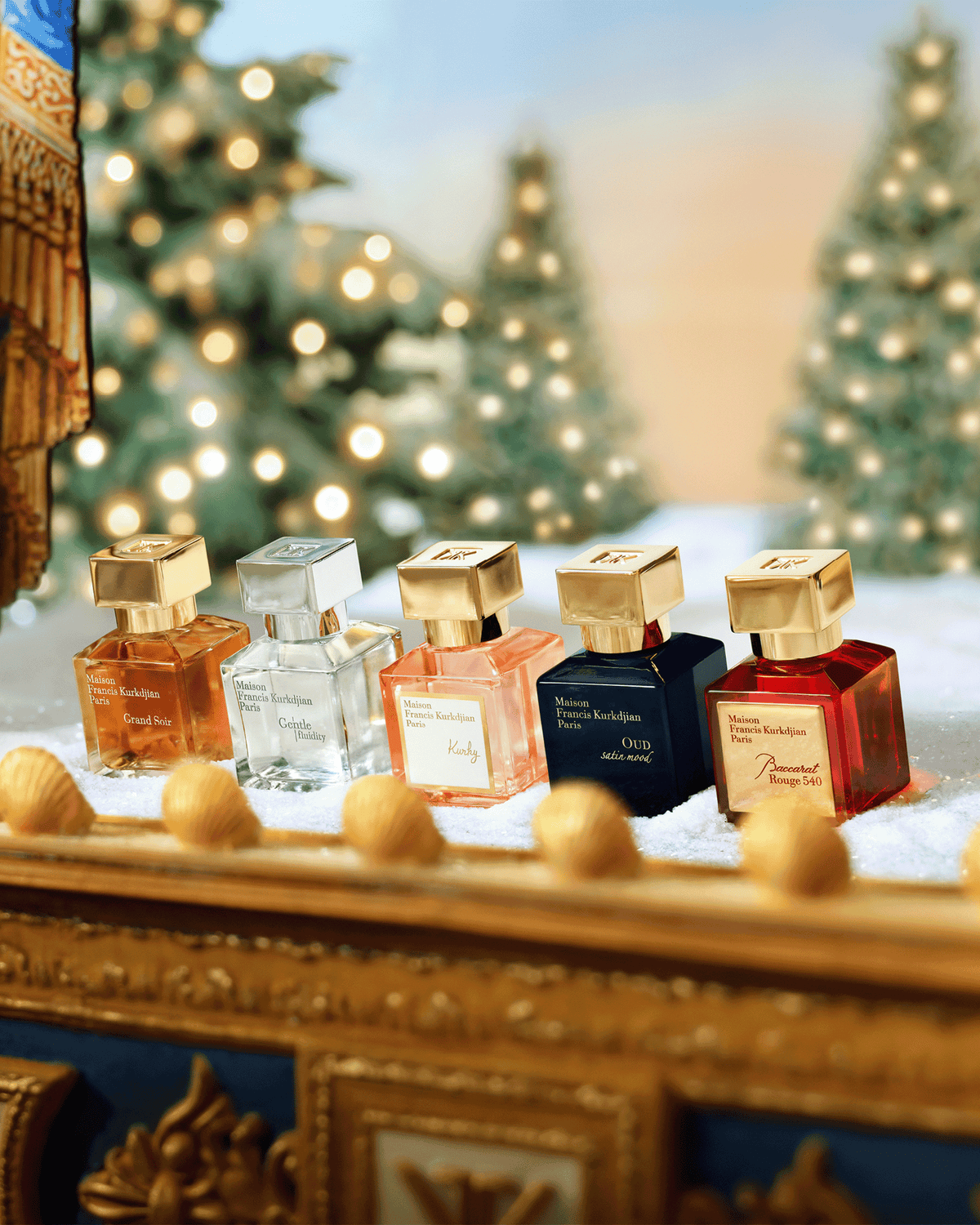 Five Maison Francis Kurkdjian miniature fragrances displayed on a snow-covered gold mantel with softly illuminated Christmas trees in the background.