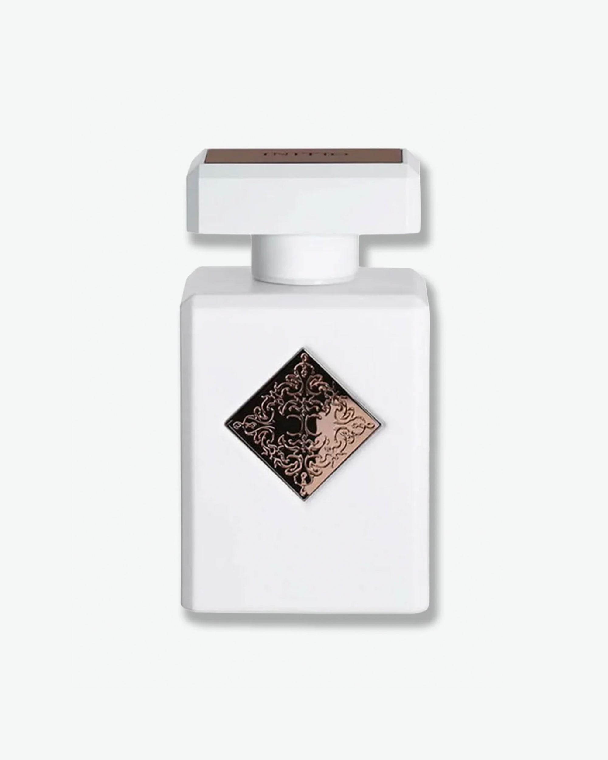 Initio Parfums Privés fragrance in a clean matte white square bottle, featuring a centered rose gold or bronze diamond badge with an intricate engraved filigree emblem, isolated on a light grey background.