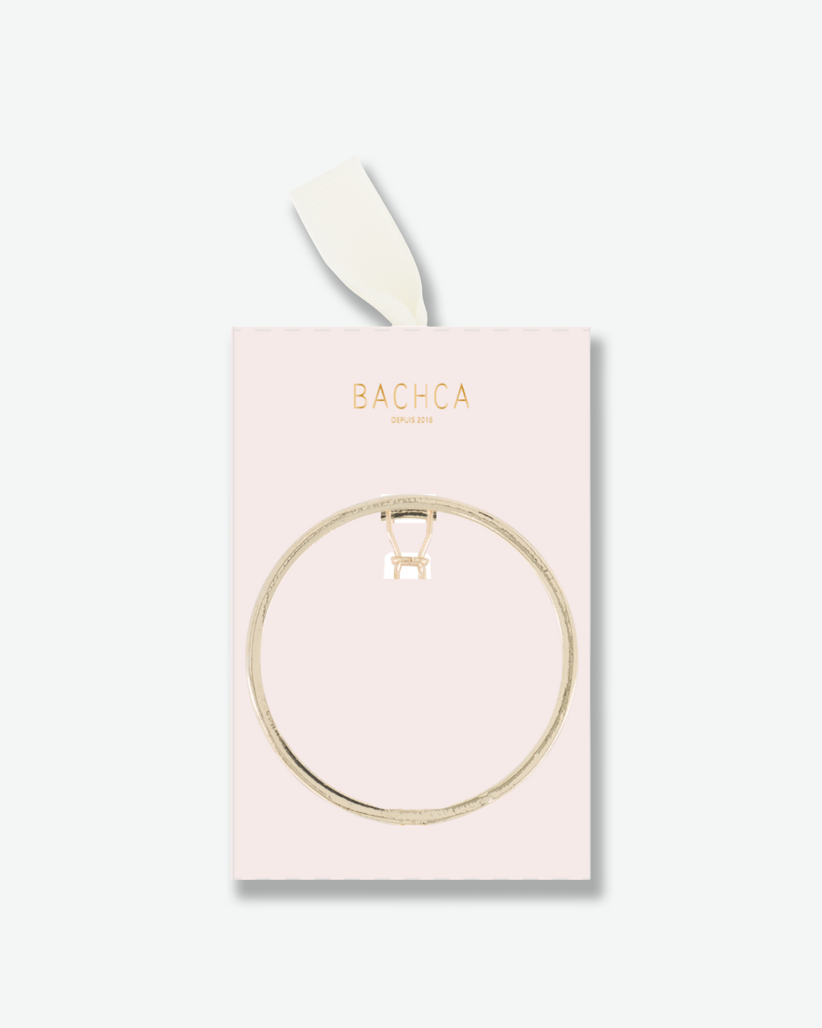 Circular gold-tone Bachca hair pin displayed on a pale pink presentation card with gold lettering and an ivory ribbon.