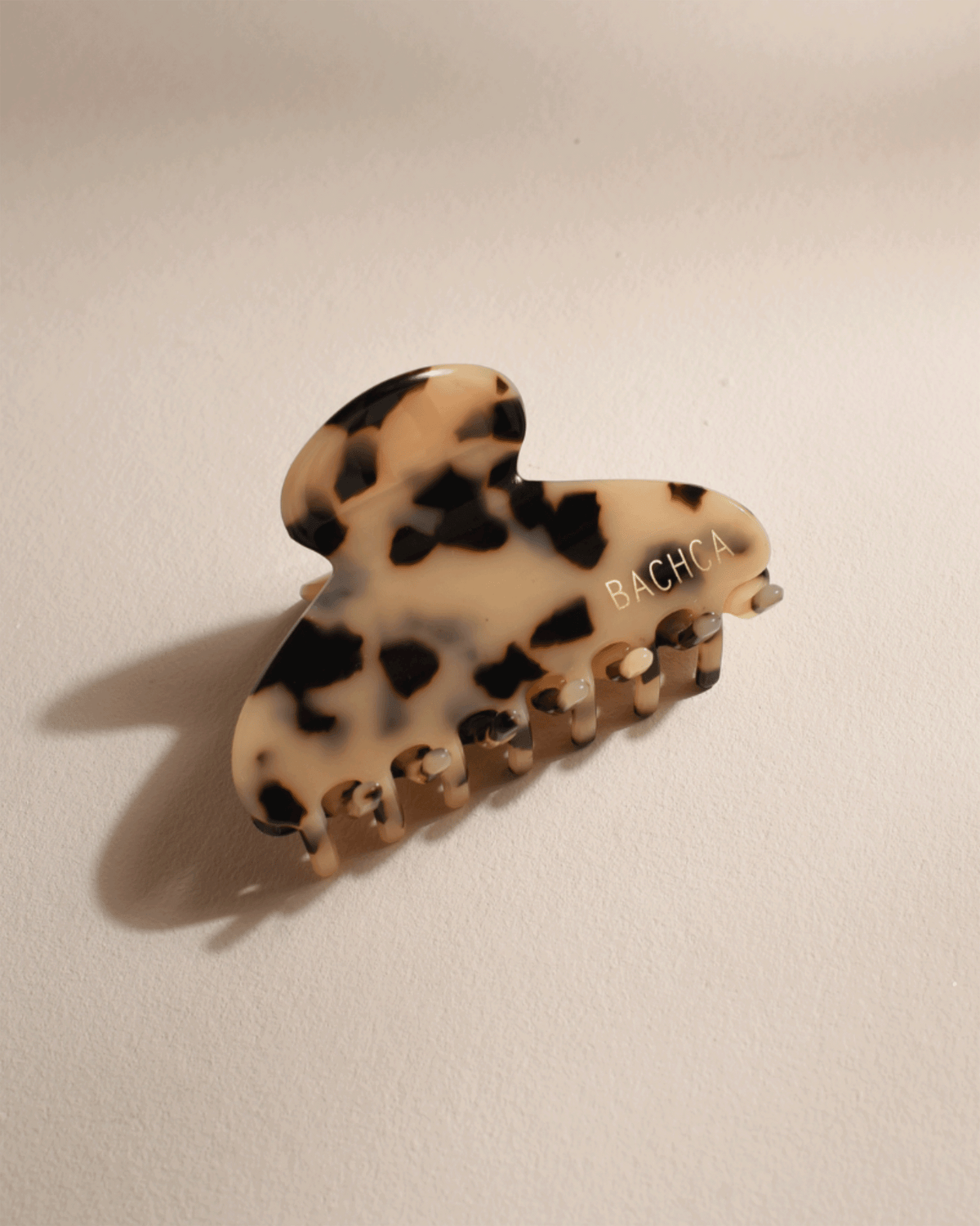 Glossy tortoiseshell Bachca Paris hair claw clip with gold logo lettering, displayed at an angle on a warm beige surface with soft shadows.