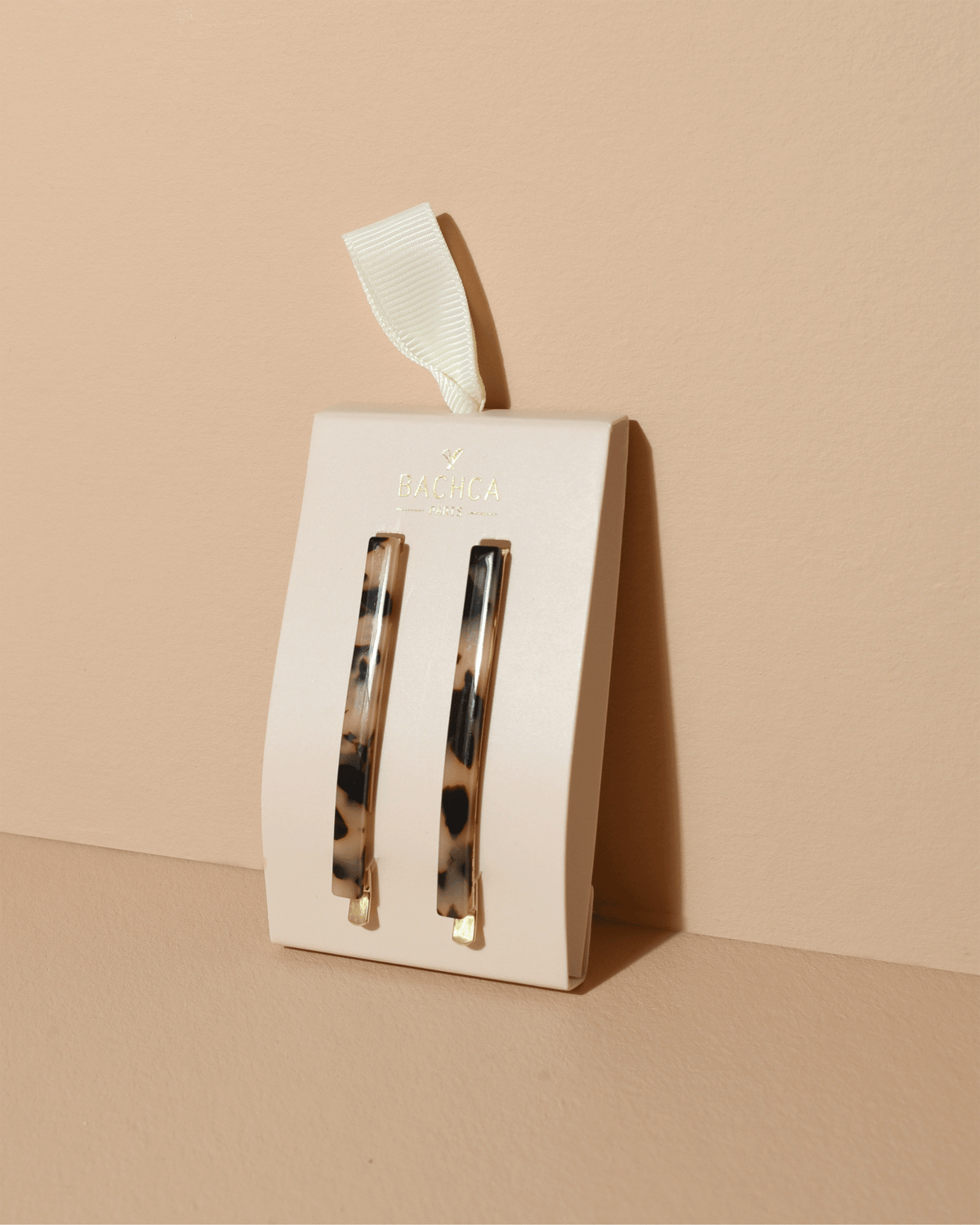 Two tortoiseshell hair clips displayed on a cream Bachca Paris presentation card with gold lettering and an ivory ribbon, against a warm beige background.