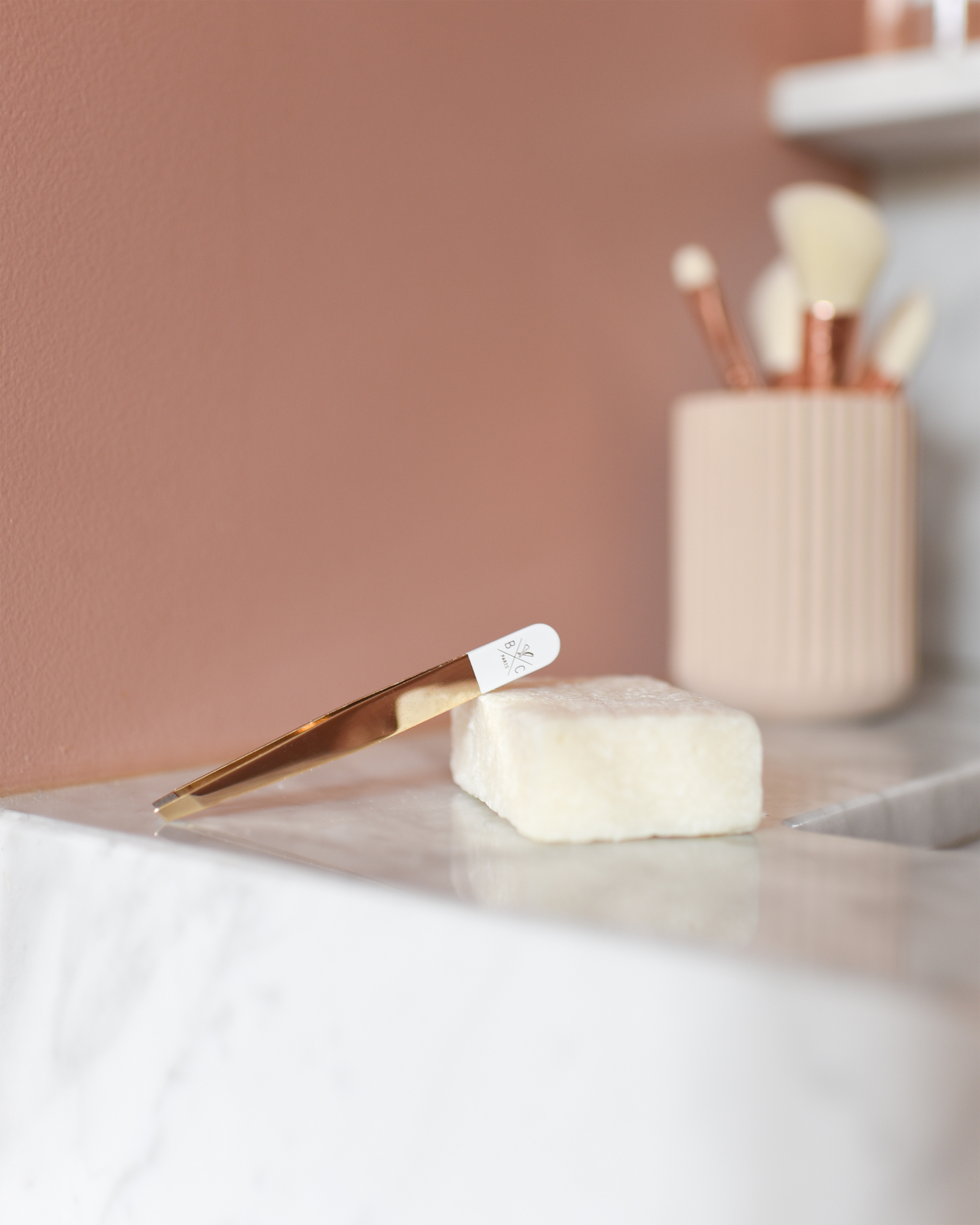 Luxury gold and white slanted tweezers resting on a bar of soap in a modern bathroom setting.