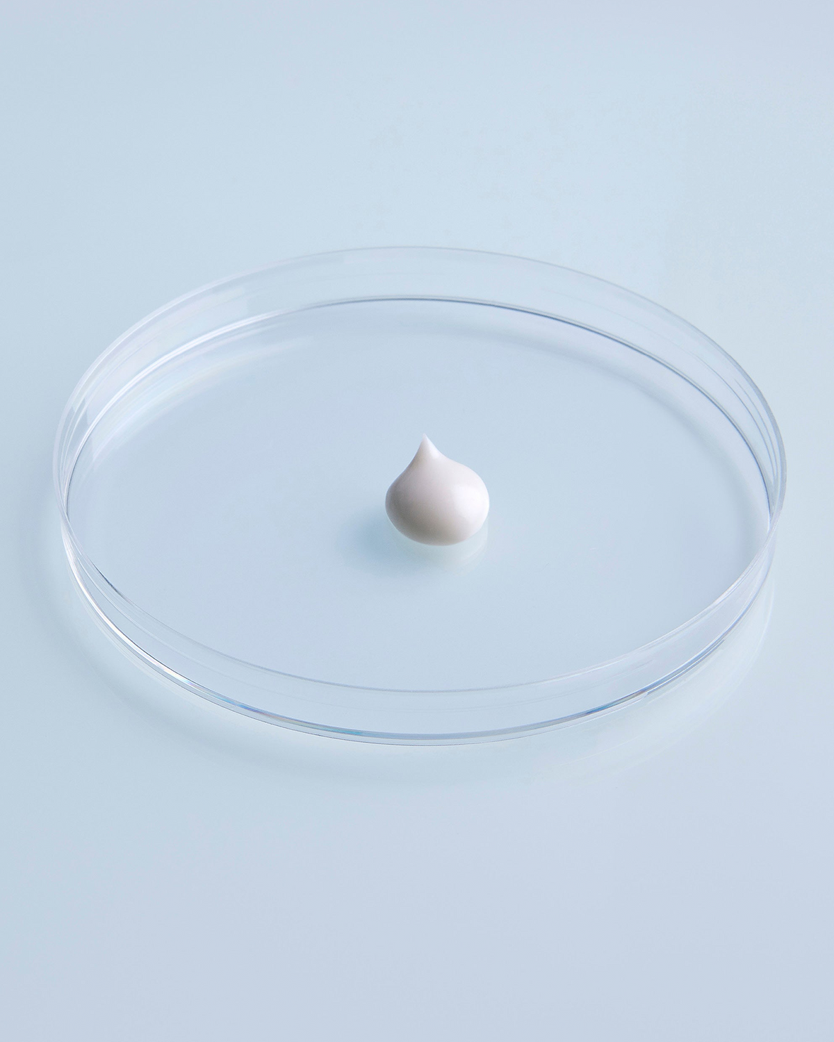 A small white cream dollop sits centered in a clear round petri dish against a pale blue background.