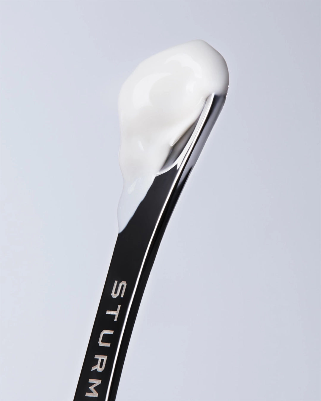 Close-up texture shot of a dollop of smooth, rich white eye cream resting on the tip of a polished black skincare spatula embossed with the brand name "STURM" in silver.