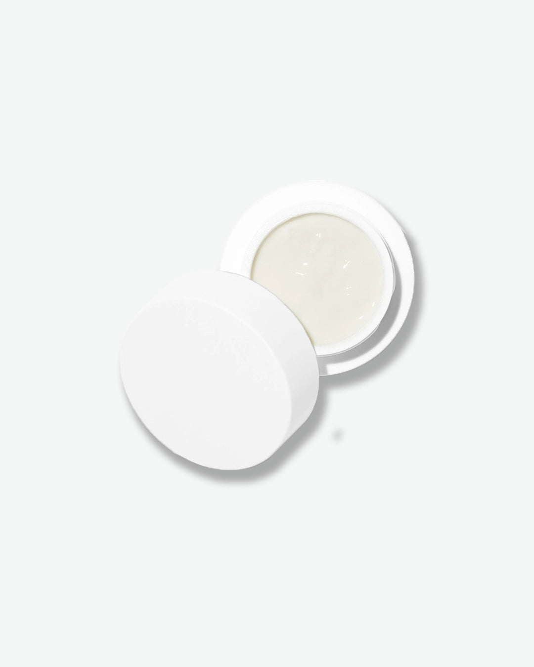An overhead, top-down view of an open Dr. Barbara Sturm Eye Cream jar against a plain gray background. The smooth white cap is placed slightly askew next to the open jar, revealing the smooth, opaque white cream inside.