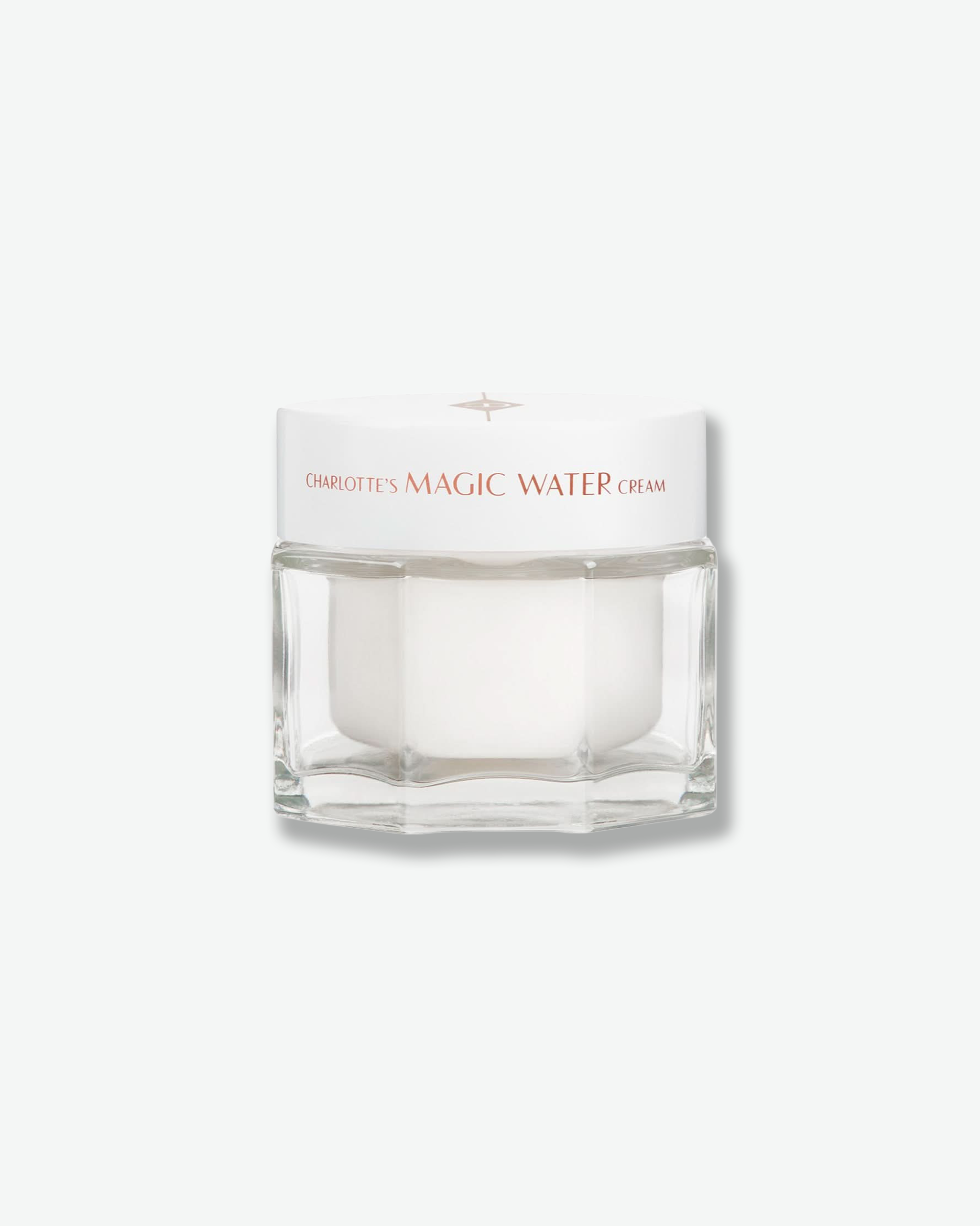 Front view of a Charlotte’s Magic Water Cream jar against a plain gray background. The product is housed in a clear, multi-faceted geometric glass jar with an opaque white inner pod, topped with a clean white screw-on lid featuring the product name in rose gold text.