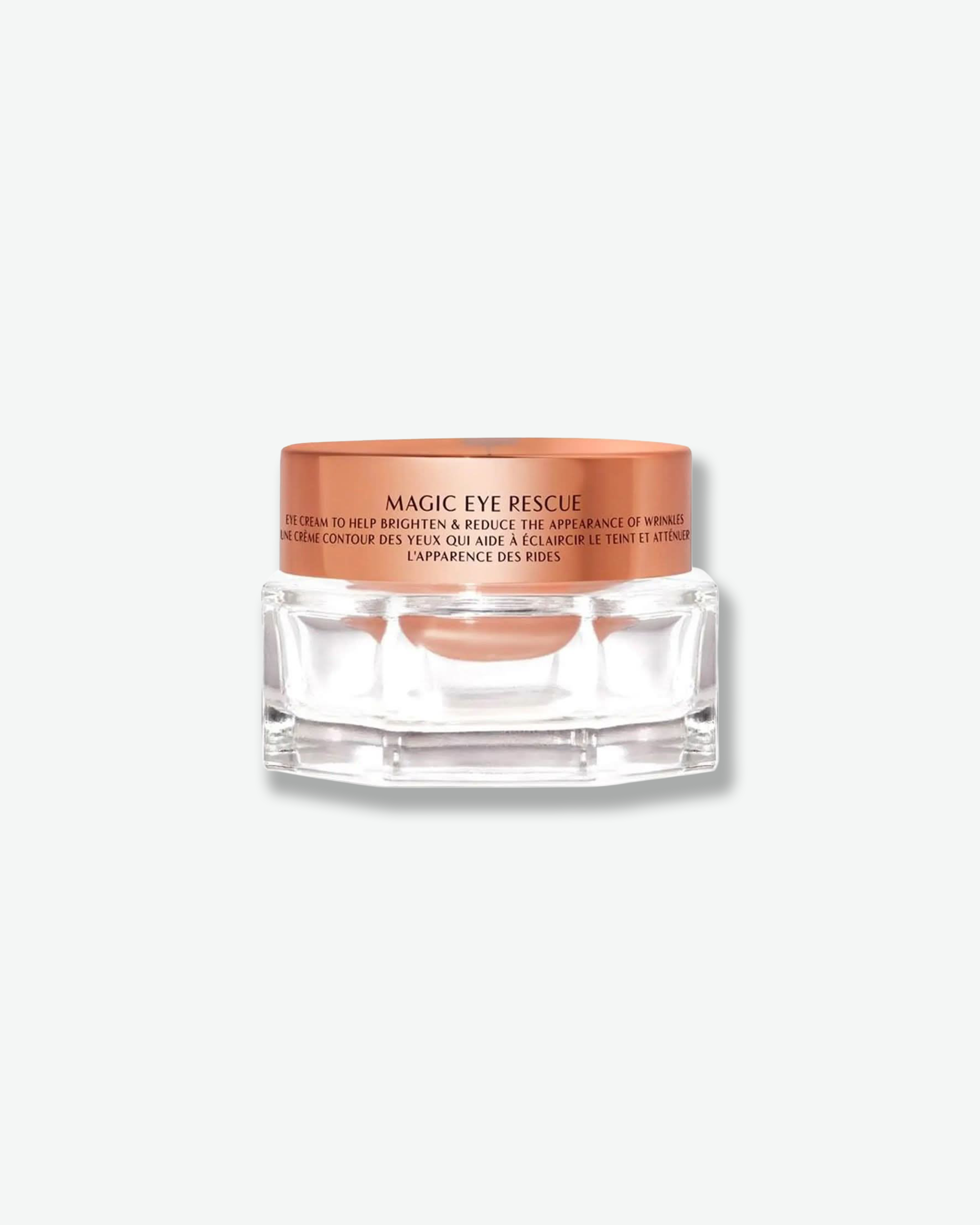 Front view of a Charlotte Tilbury Magic Eye Rescue cream jar against a plain gray background. The premium design consists of a clear, multi-faceted geometric glass jar showing the peach-tinted eye cream inside, capped with a reflective, metallic rose gold lid printed with black product details.