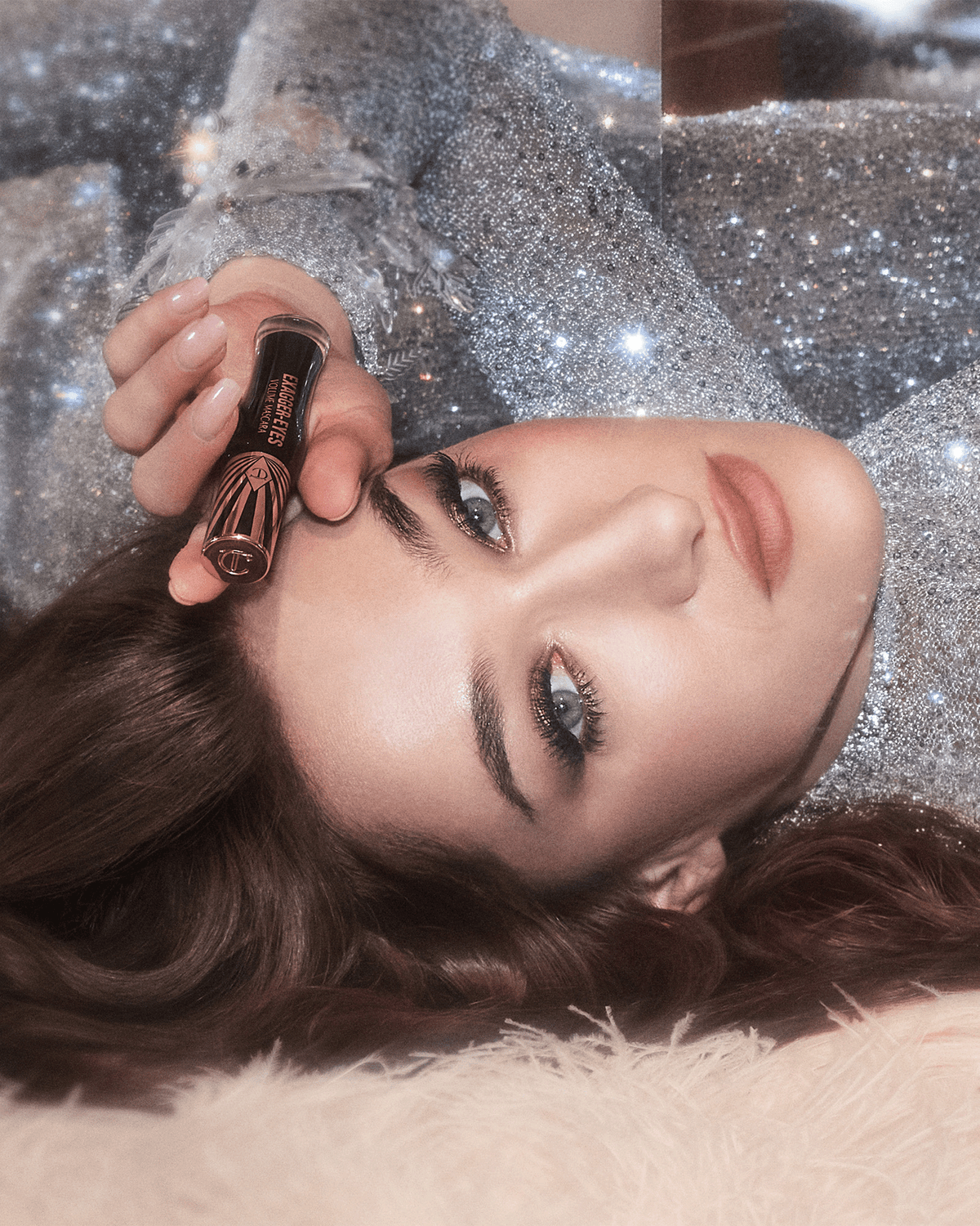 Model holding Charlotte Tilbury Exagger-Eyes Volume Mascara against a sparkling silver backdrop.
