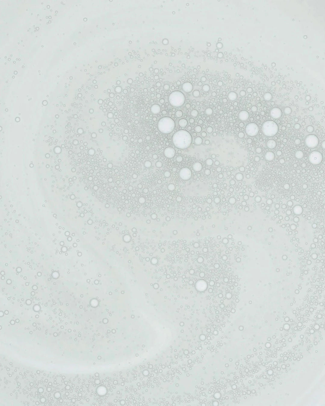 An abstract close-up of a milky skincare formula featuring hundreds of suspended microbubbles. The lightweight emulsion is shown in a swirling pattern against a pale background, highlighting its smooth texture and blend of oil-and-water components.