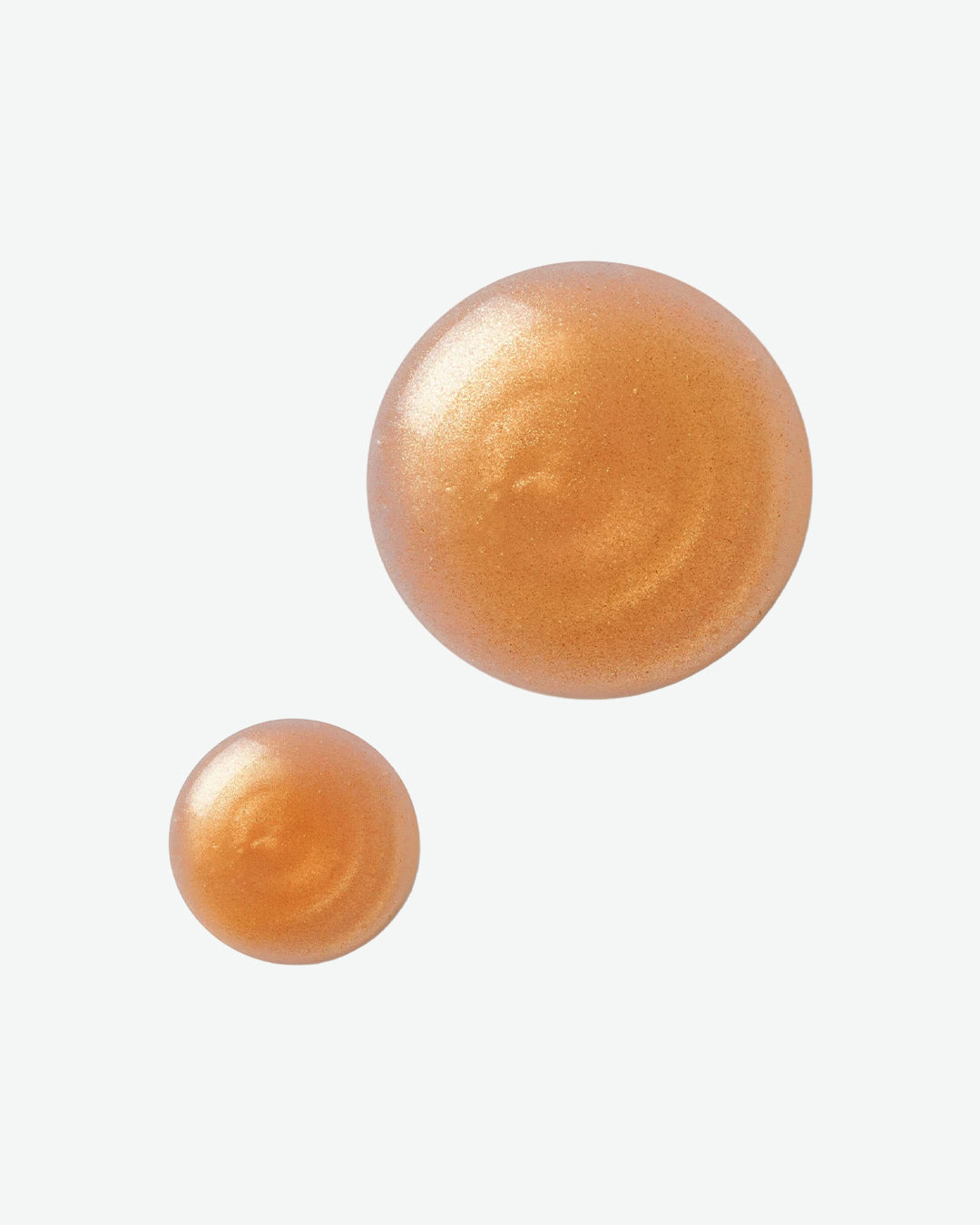 Two swatches of a shimmering golden skincare or makeup formula are displayed against a light gray background. The pearlescent product appears in circular droplets of different sizes, showcasing its luminous finish, smooth texture, and radiant glow.