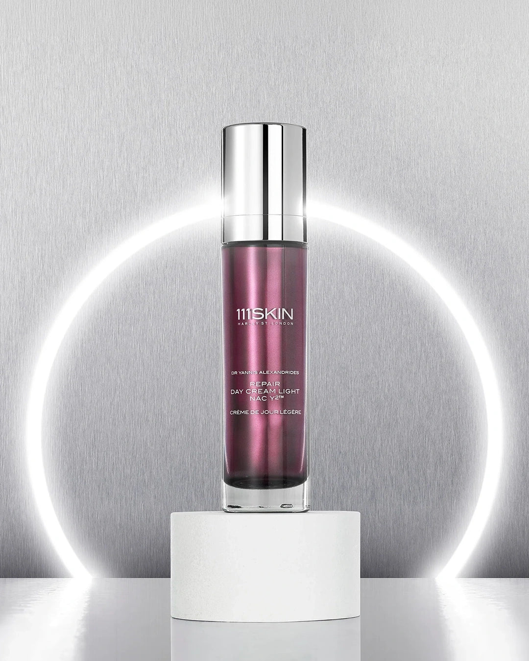 Metallic maroon bottle of 111Skin Repair Day Cream Light NAC Y2 with silver cap centered on a white pedestal against a silver backdrop with a white ring light illuminating it from behind
