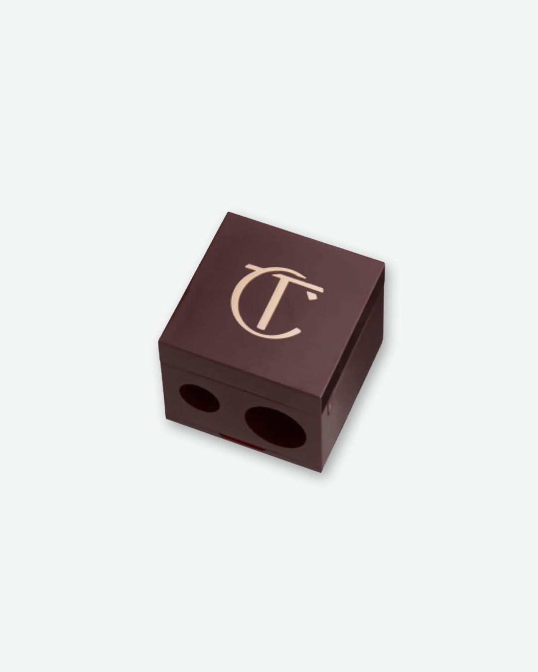 An angled product shot of a Charlotte Tilbury dual-size eyeliner and lip pencil sharpener, centered against a plain light gray background. The square, cube-shaped sharpener is a deep signature night-crimson color. Its top lid features a gold "CT" monogram logo, and the front face displays two sharpening holes of different sizes to fit standard and jumbo makeup pencils.