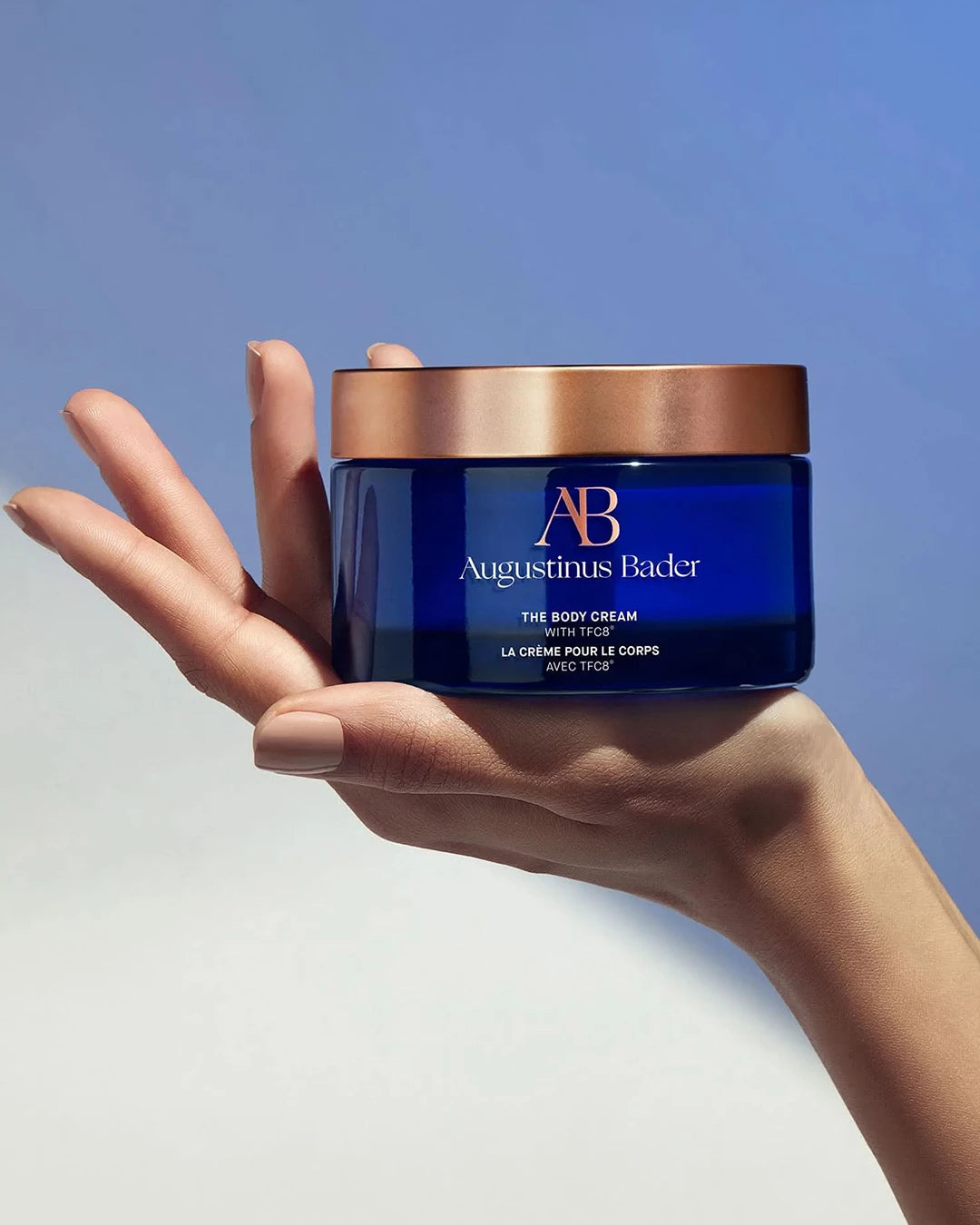 Hand holding a blue jar of Augustinus Bader cream against a light background
