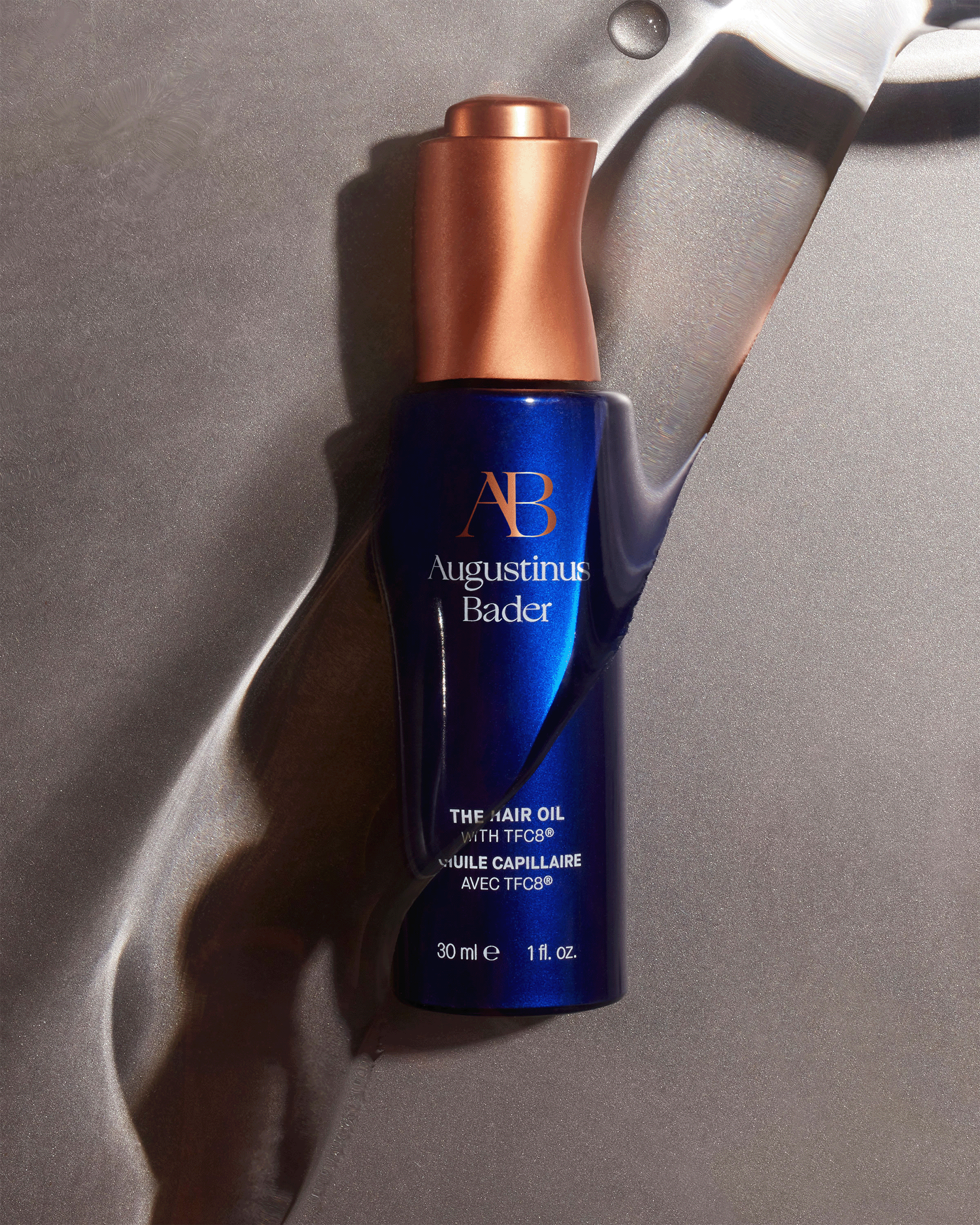 An Augustinus Bader hair oil bottle lying flat against a wavy, shimmering metallic silver texture background.