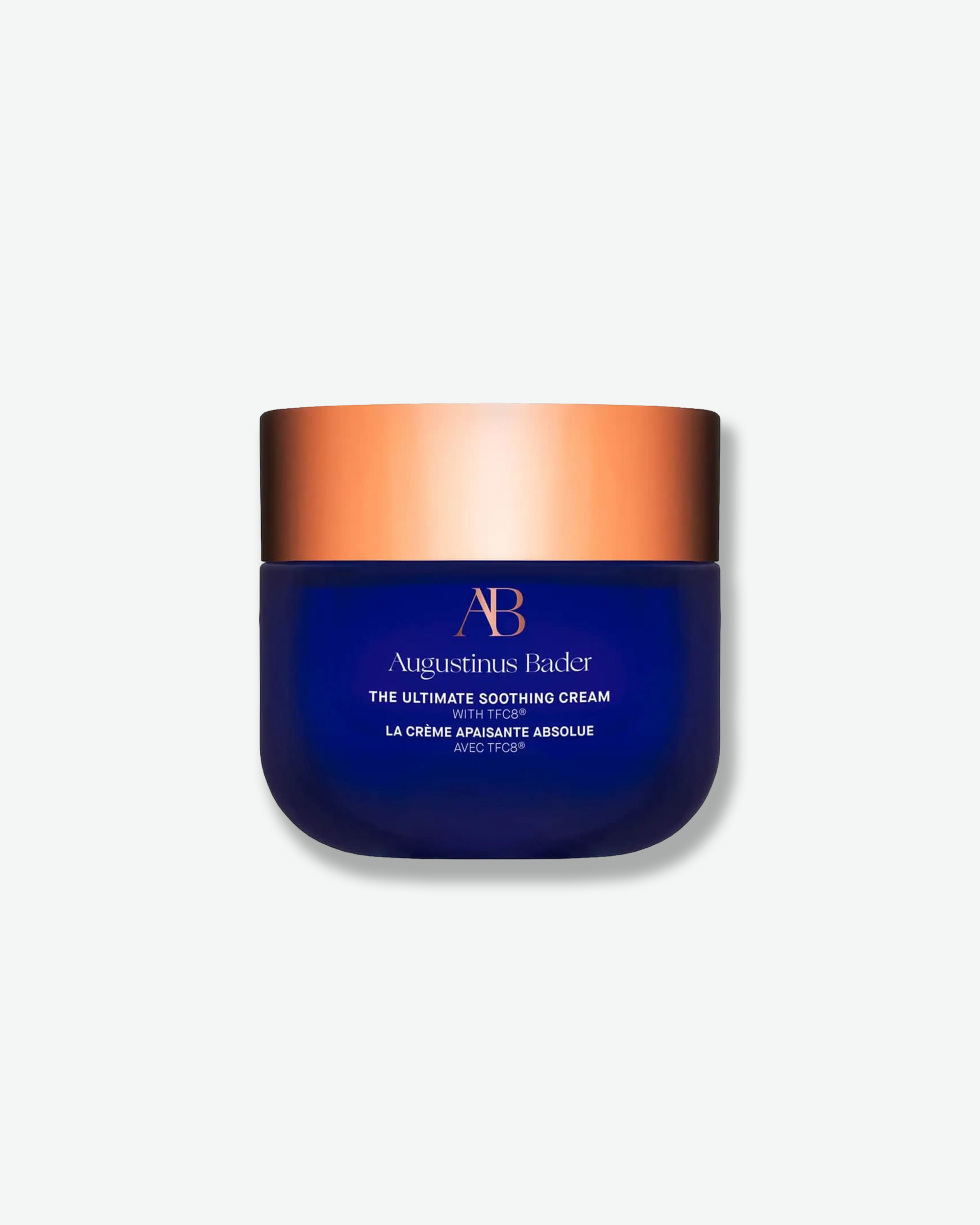 Front view of an Augustinus Bader "The Ultimate Soothing Cream" jar against a plain gray background. The design features a smooth, deep royal blue base with rounded bottom corners, showcasing copper-gold logo accents and white text branding, topped with a wide, metallic copper-gold cap.