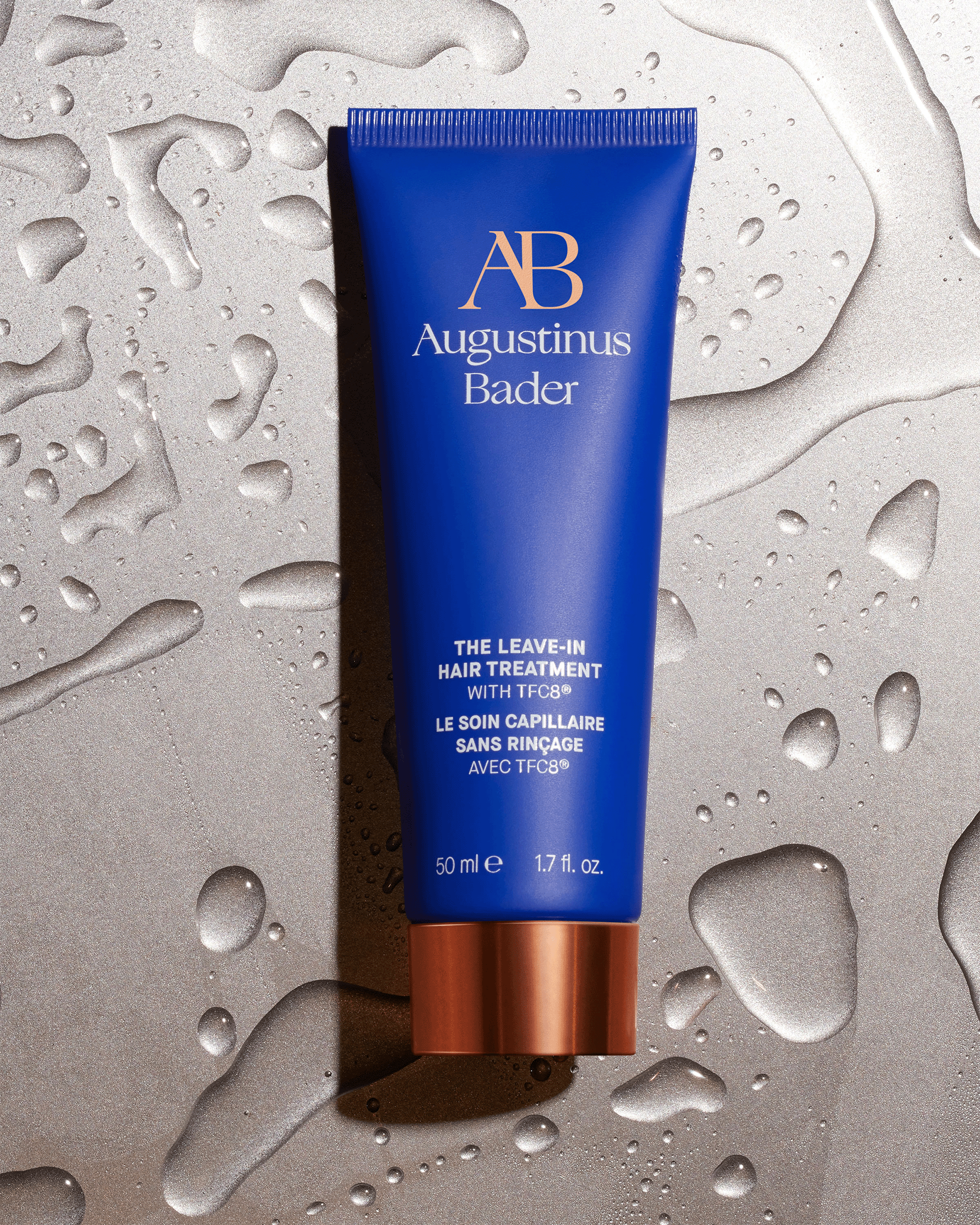 An Augustinus Bader blue leave-in hair treatment tube lying flat on a shimmering, textured metallic gray surface covered in water droplets.