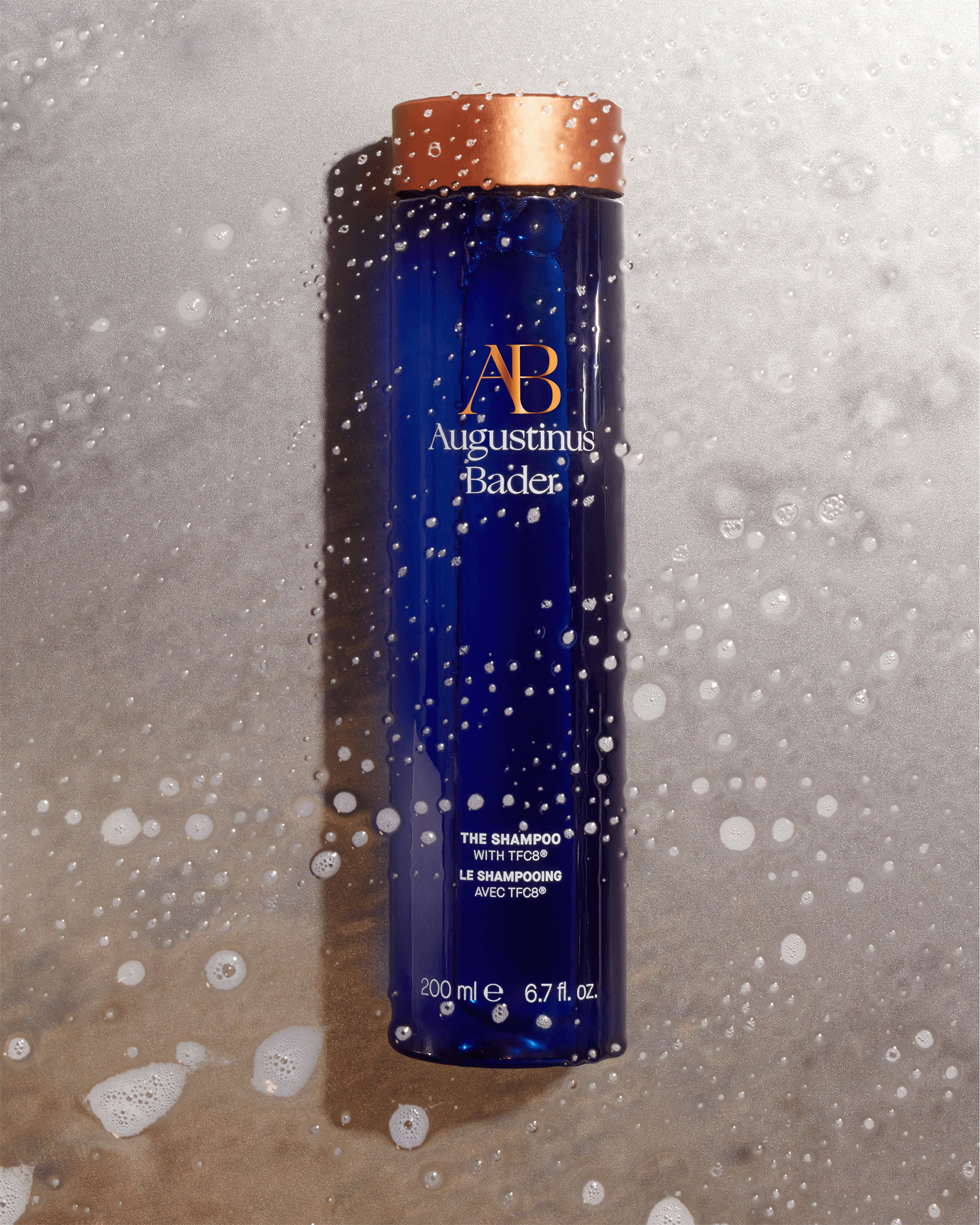 An Augustinus Bader blue shampoo bottle lying flat on a textured gray stone surface covered in water droplets and soapy white lather.