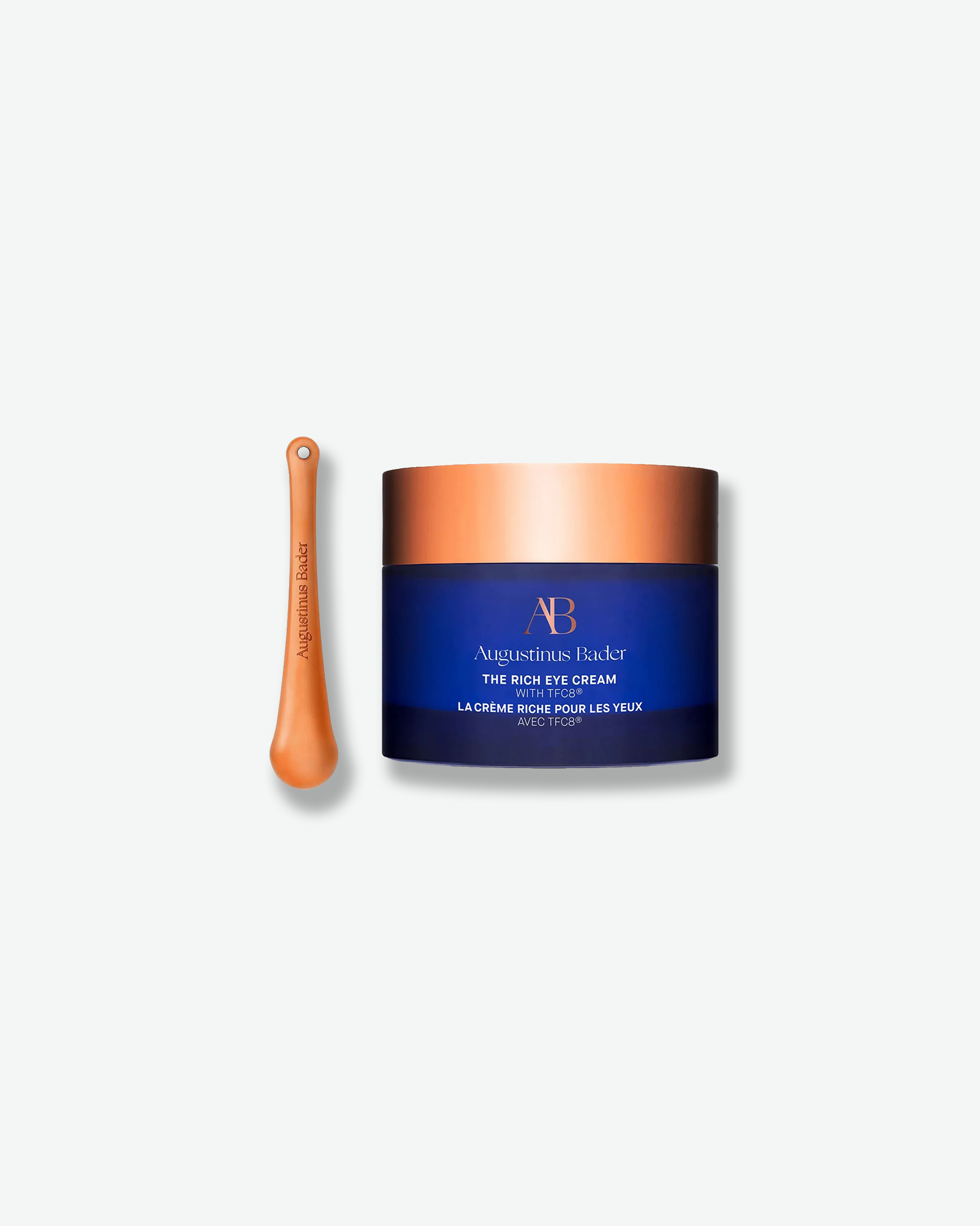 Product shot featuring an Augustinus Bader "The Rich Eye Cream" jar standing next to its application tool against a plain gray background. The royal blue cylindrical jar with a wide, metallic copper-gold lid is positioned beside a slender, matching copper-gold metallic massage spoon embossed with the brand name.