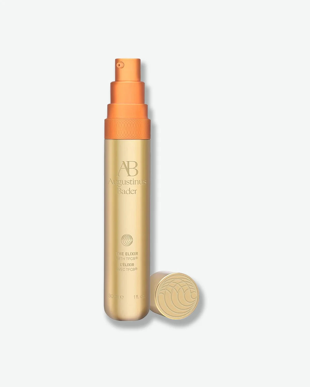 Skinny gold Augustinus Bader The Elixer vial, with an orange push pump, and cap off and next to it, centered against a light grey background.