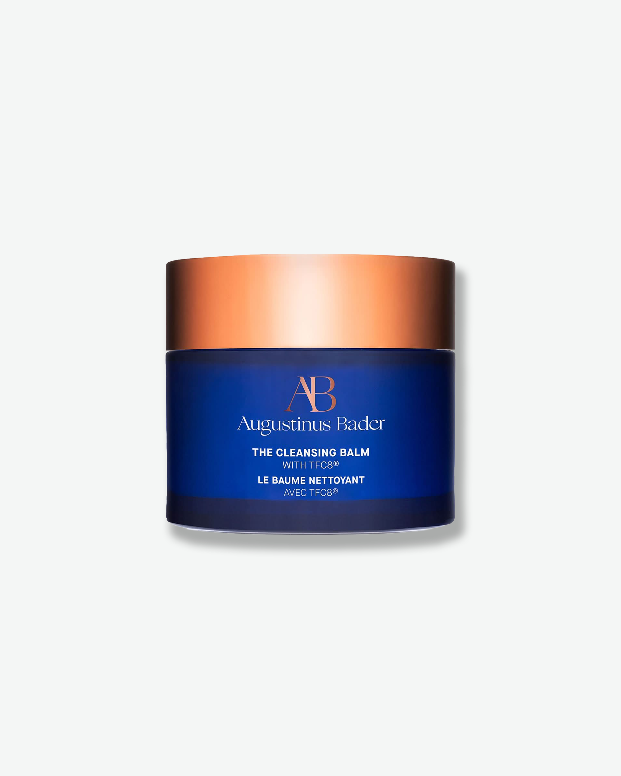 Front view of an Augustinus Bader "The Cleansing Balm" jar centered against a plain gray background. The cylindrical container features a dark royal blue body with the brand's monogram logo in copper-gold and product description in white, topped with a flat, metallic copper-gold screw lid.