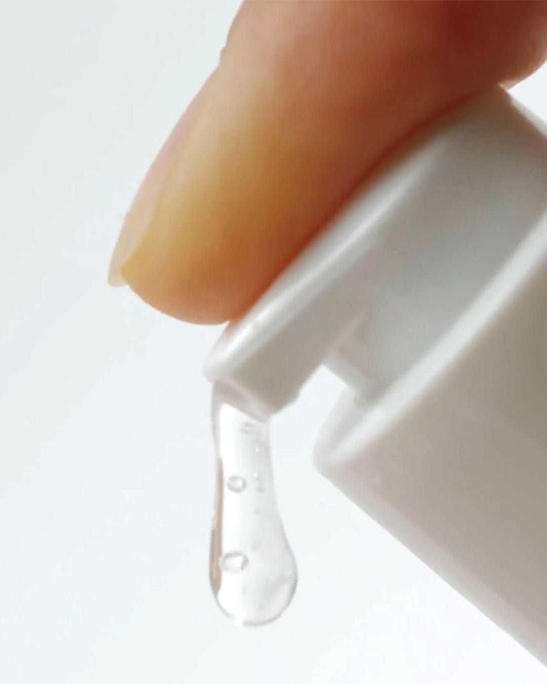Close-up of a hand holding a white bottle with a dropper, releasing clear liquid onto a light gray background.