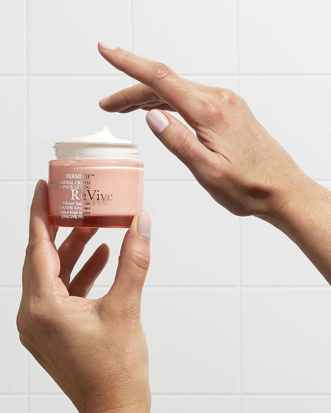 Person holding a pink jar of Revive cream with a white tile background