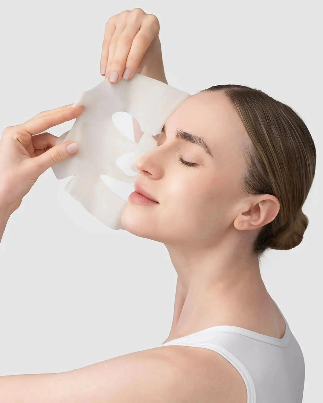 Woman applying a white sheet mask to her face against a light gray background